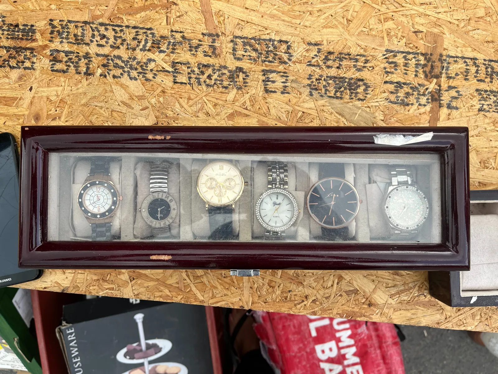 Six wrist watches in a wooden box with a glass top. One of the watches points at 33 minutes past 12, another one, with three extra dials, points at 27 minutes past 3.  There are two watches with quite a few diamonds. One of them points at 36 minutes past 6, the other at 6 minutes past 7. One watch is displayed upside down. It has the word Kipling printed in large type on its face, once from left to right, and once mirrored from right to left. This watch points at 8 o’clock. And finally there’s this one more watch that points at 21 minutes to 12.