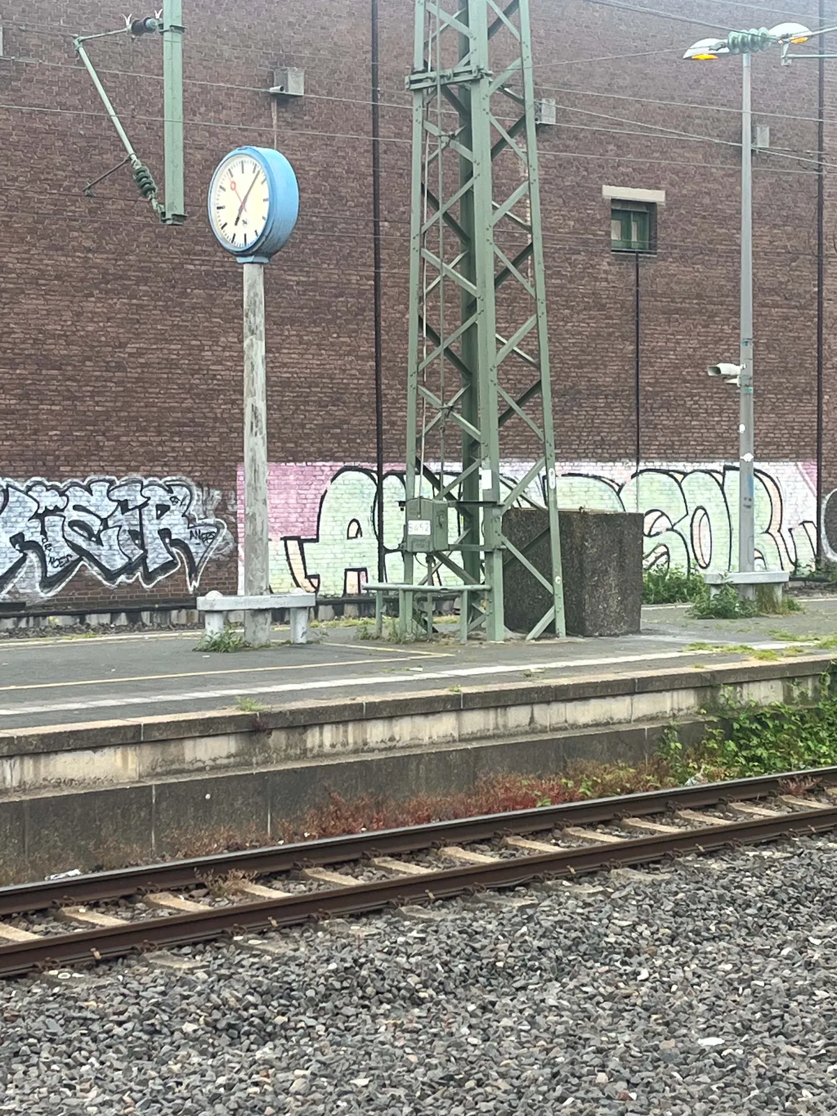 Looking across the tracks, at a clock on a pole on a train platform. Behind the clock there’s a very large brown brick wall. The clock points at 7 minutes past seven.