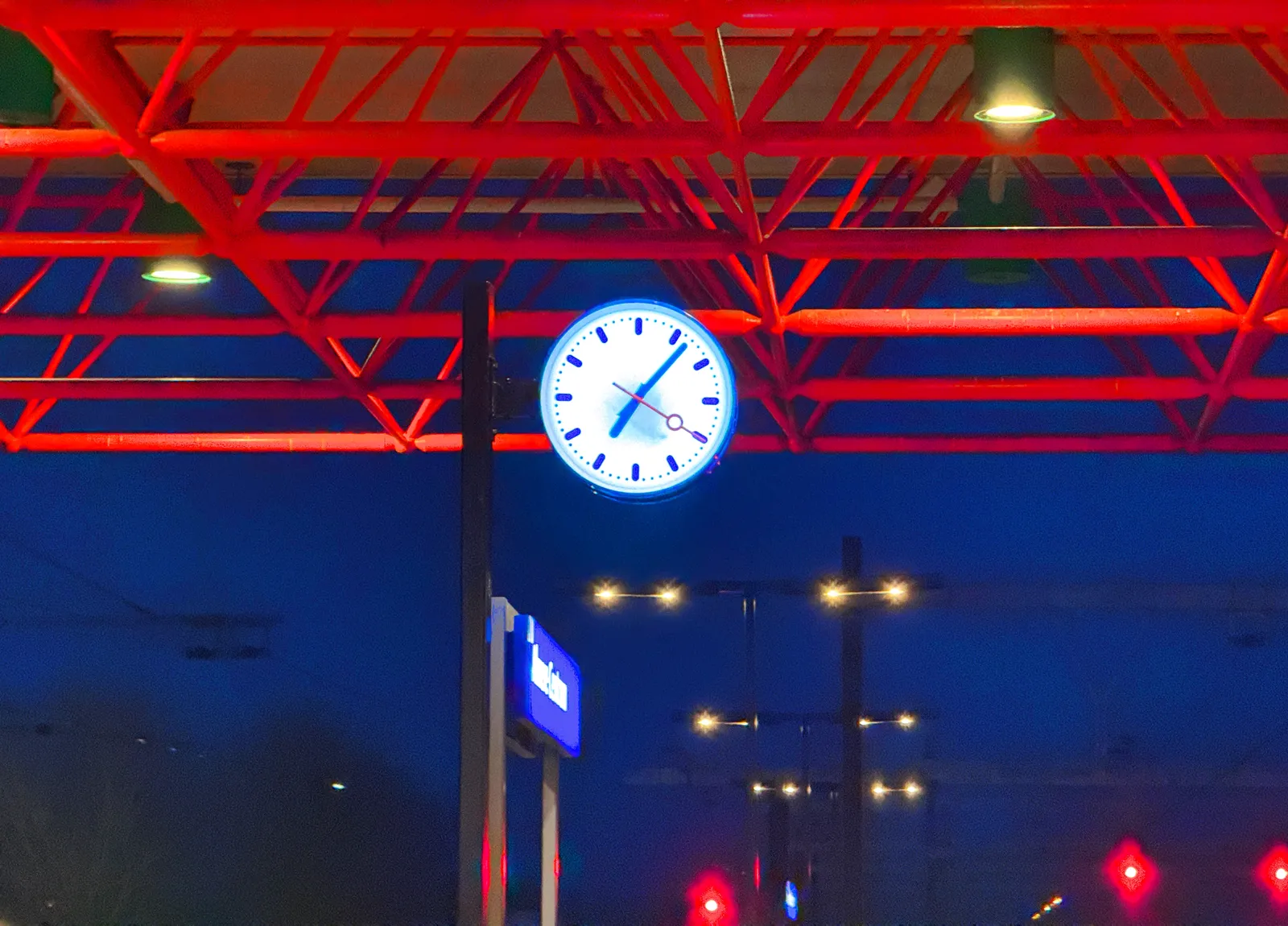The bright red construction of the roof of a Dutch train station fills the upper part of the image. In the center there’s a lit clock on a pole which points at 7 minutes past seven.