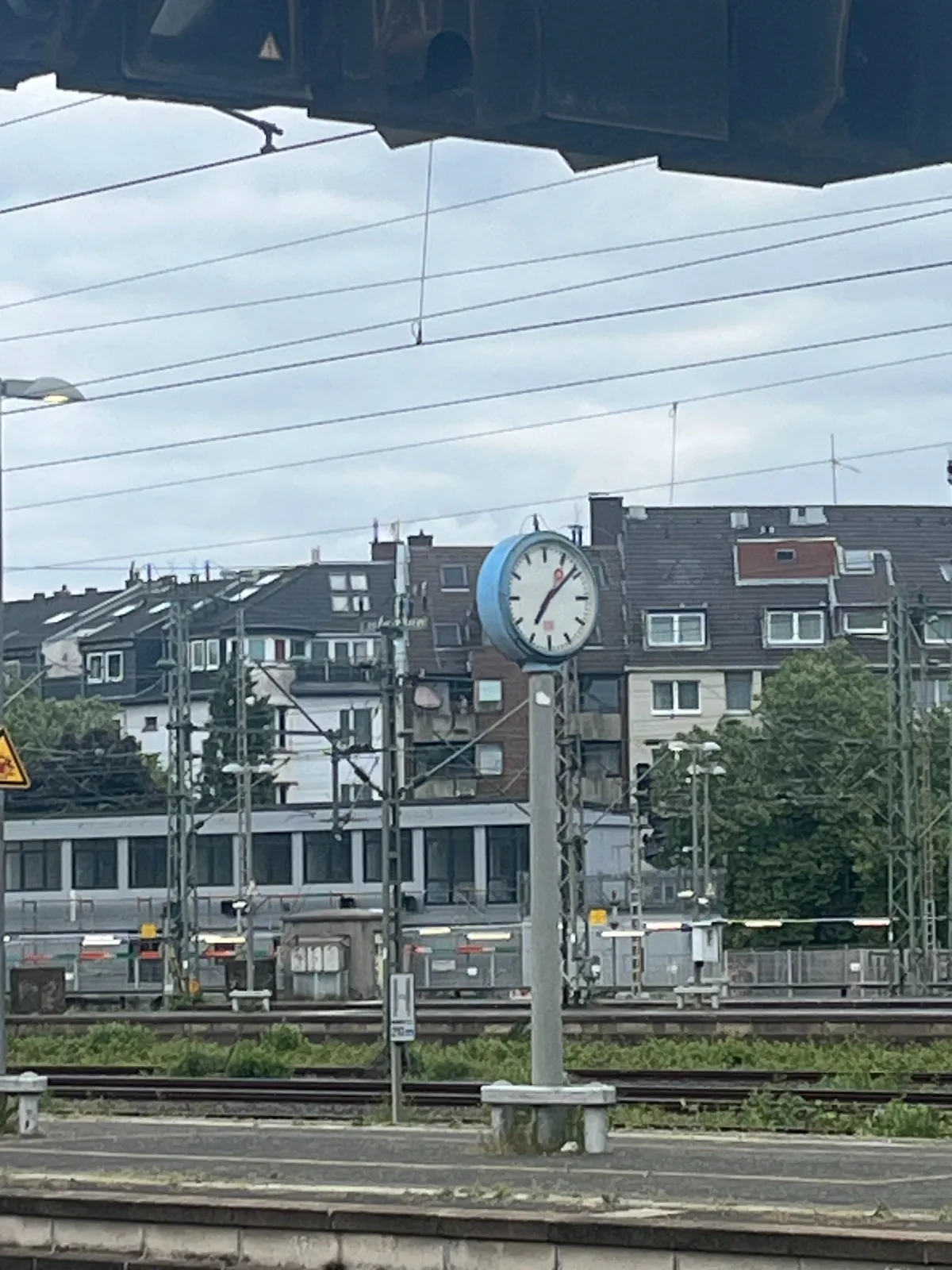 There’s a clock on a pole in the distance. Even further behind it are some high appartment buildings. The clock points at 8 minutes past seven.