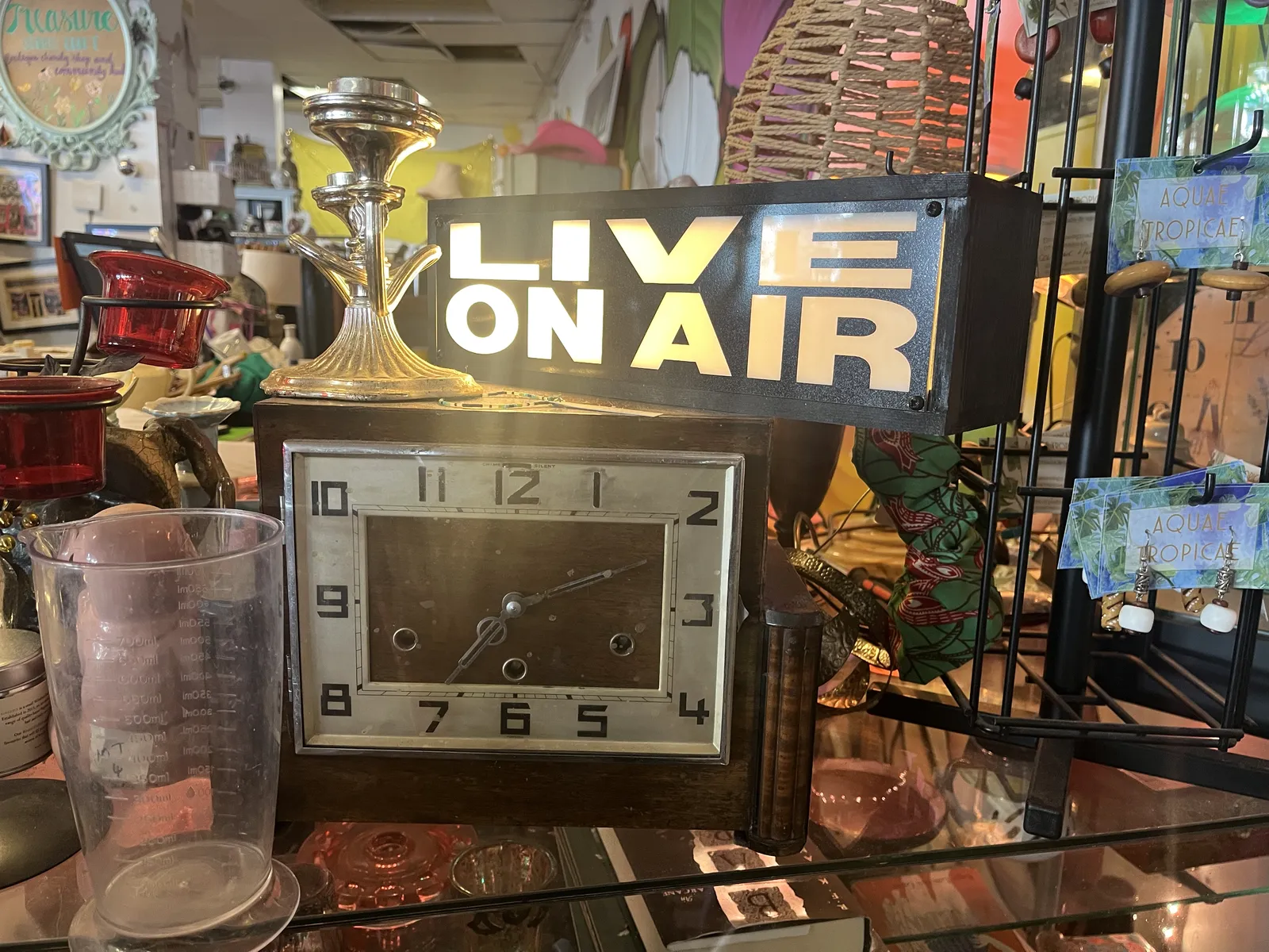 All kinds of things for sale. Among these things there’s a wooden mantel clock, with a lit sign on top of it that says Live On Air. The clock points at 11 minutes past seven.