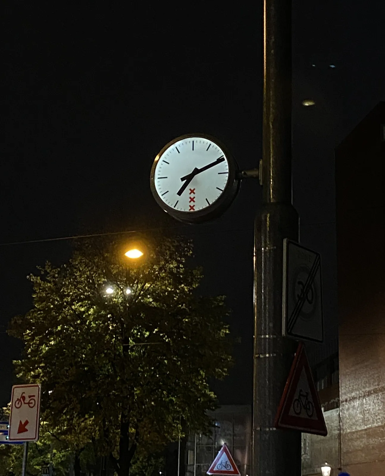 A clock in Amsterdam, attached to a street light. There are all kinds of road signs around it. The clock points at 11 past 7.