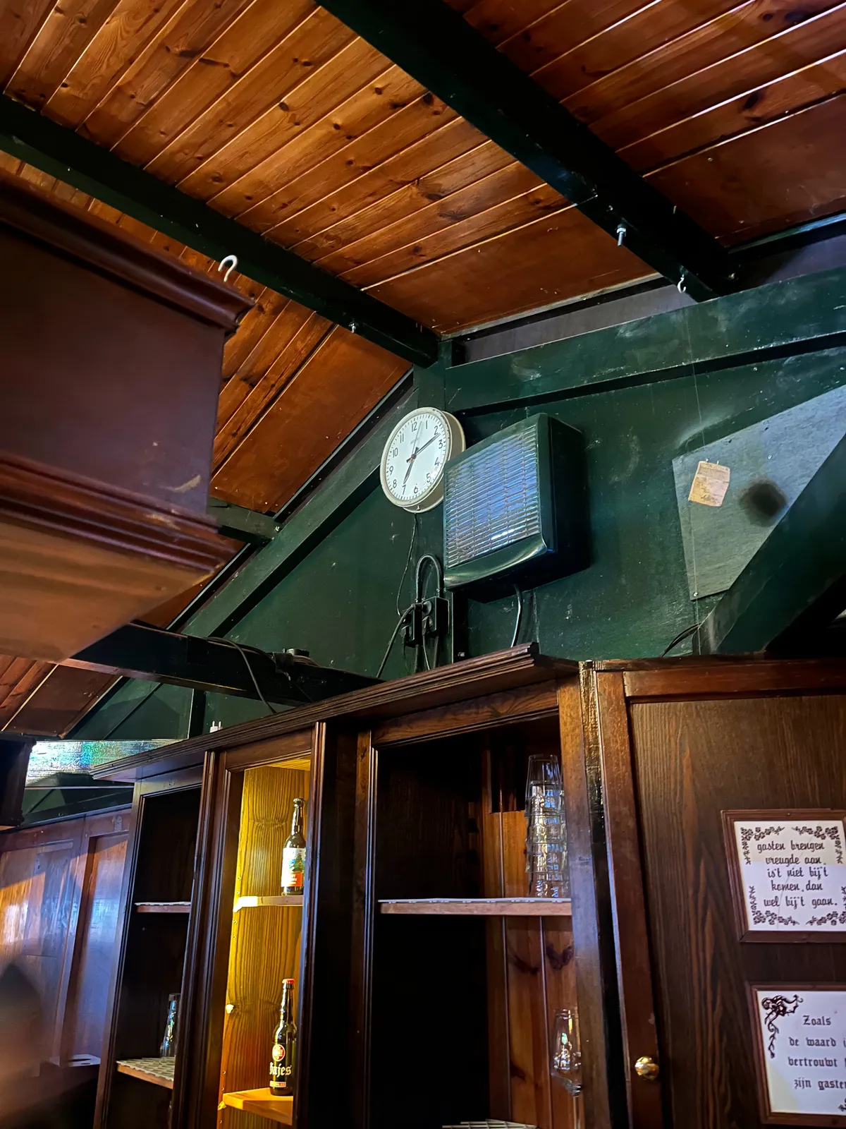 Inside a wooden bar. A dark wooden roof, and wooden cupboard, painted in dark green with a few bottles of beer in it. There are two so called wisdom tiles attached to this cupboard. One of them says, in Dutch: Guests bring joy. If not when then enter, then when they go. The other one, again in Dutch: the way the innkeaper is, is the way he trusts his guests. There’s a clock above this cupboard. It points at 12 minutes past 7