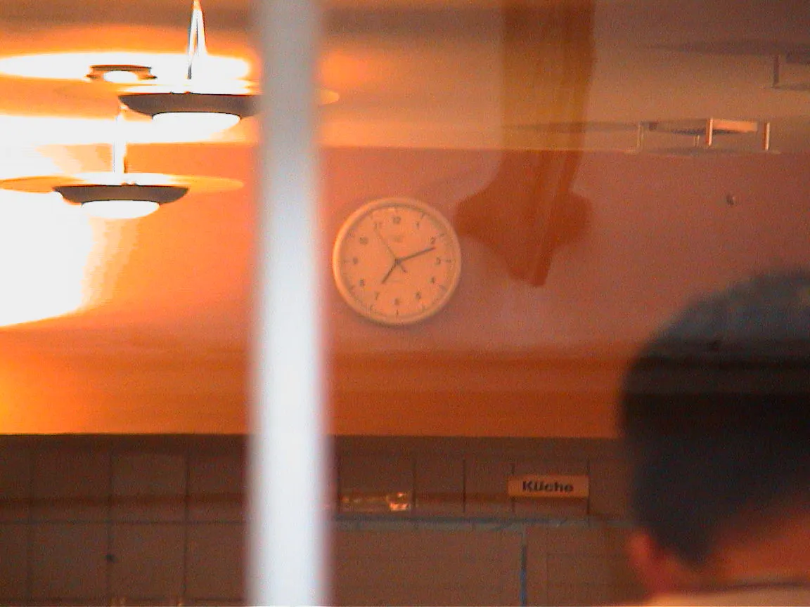 A clock seen through a window points at twelve past seven.