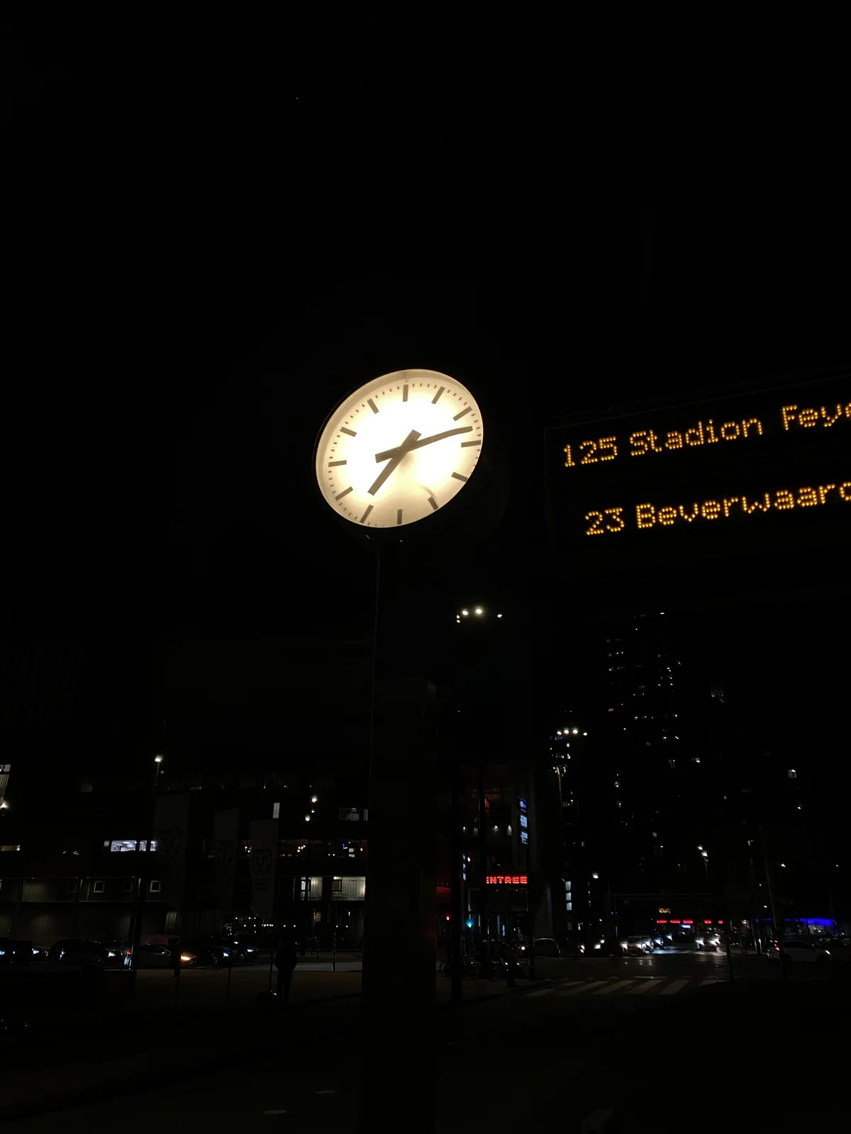 A lit bus stop sign in darkness. There are some partial destinations visible: 125 stadion Fey and 23 Beverwaar. There’s also a lit clock. It points at 13 minutes past 7.