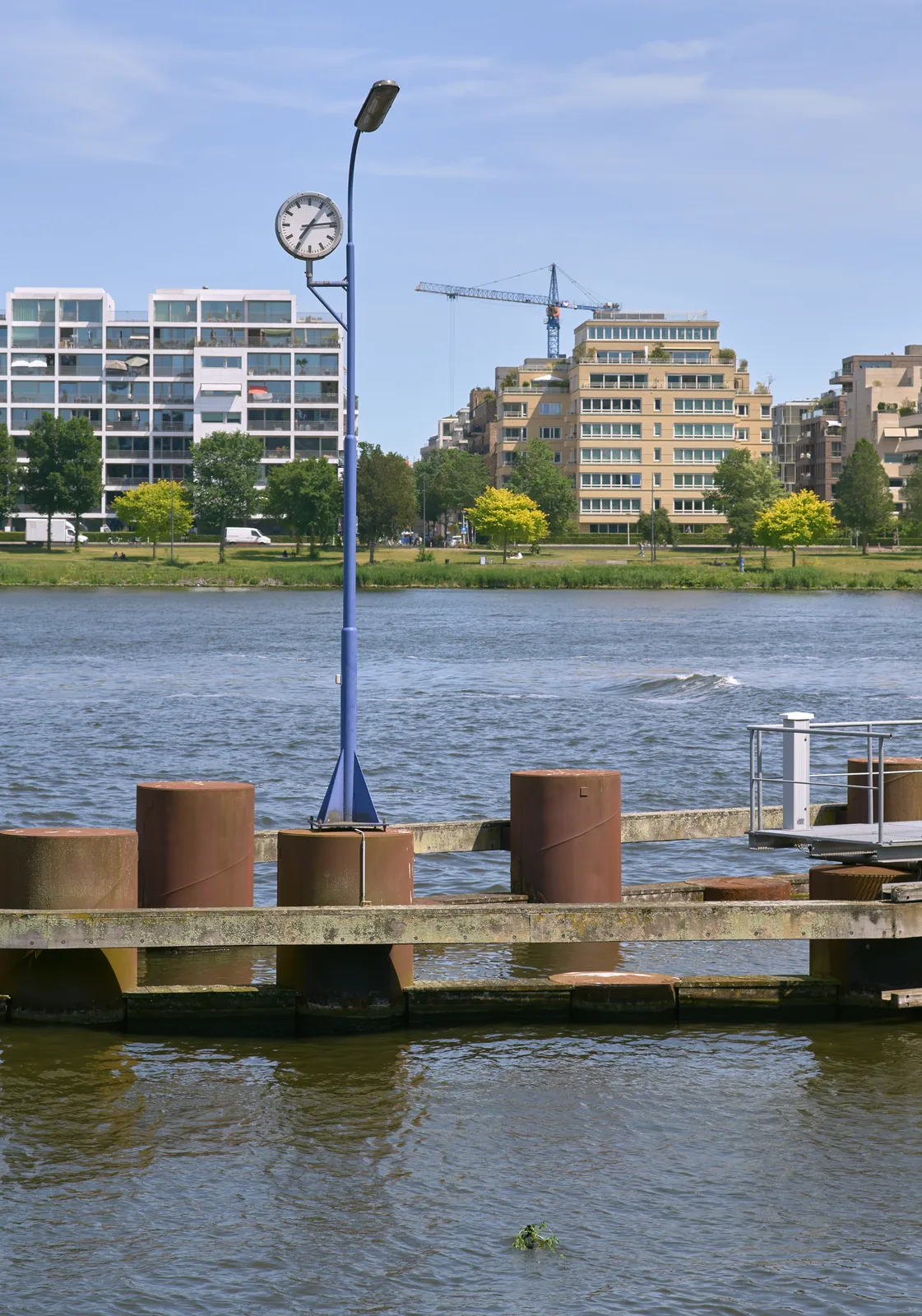On the other side of the water there are some appartment buildings. In the water there’s a part of a jetty, with a clock on a pole. This clock points at 14 minutes past seven