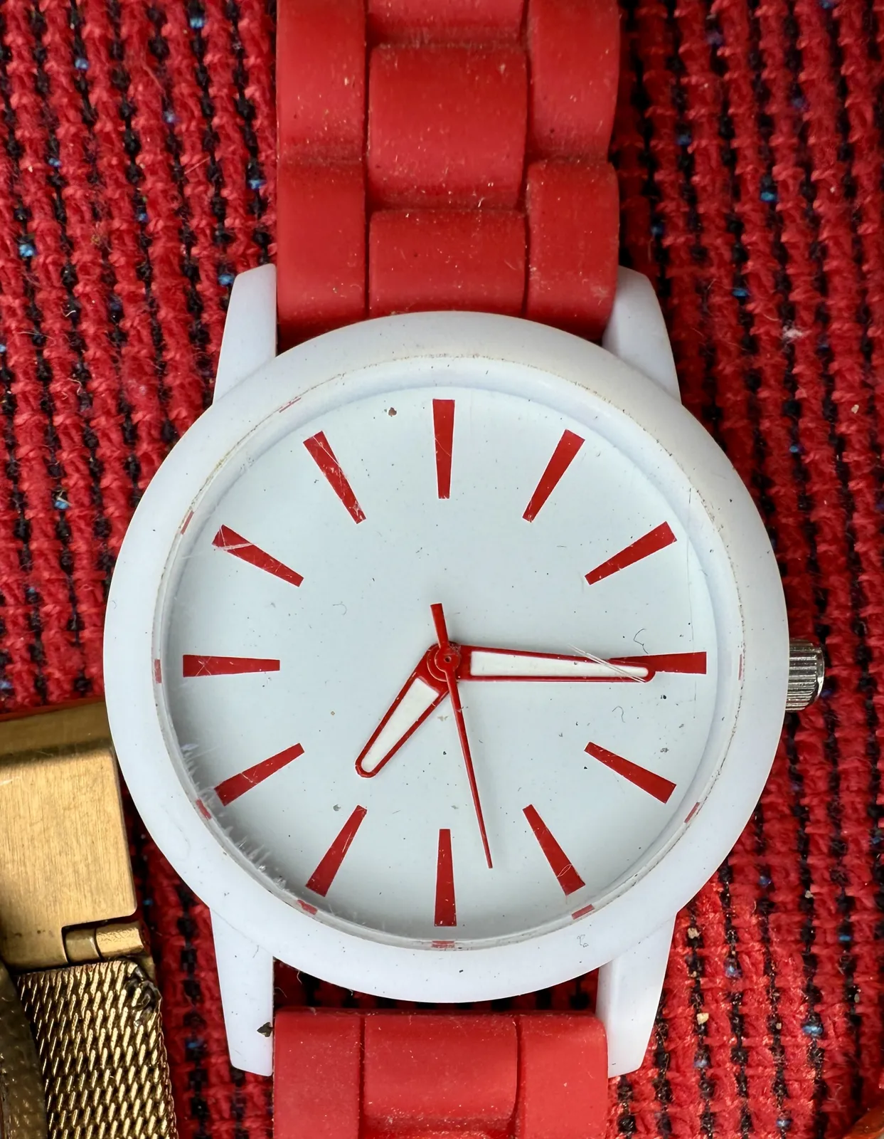 A white clock with a red strap, red hands and red indicators points on a red rug. It points at a quarter past seven.