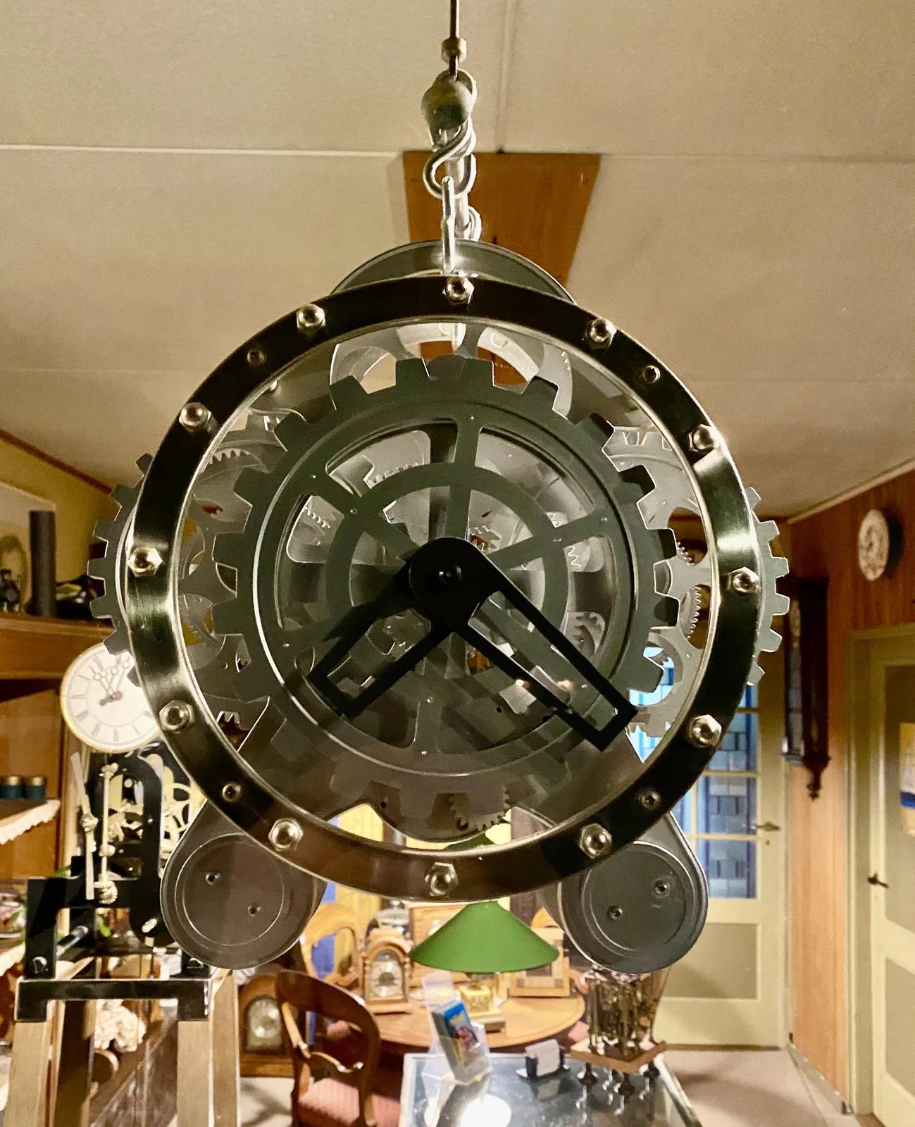 A clock hanging in front of a window. Its cogs are visible, and it has very wide hands. This makes it unclear to see what time it is exactly. So lets just assume it points at approximately 21 minutes past 7.