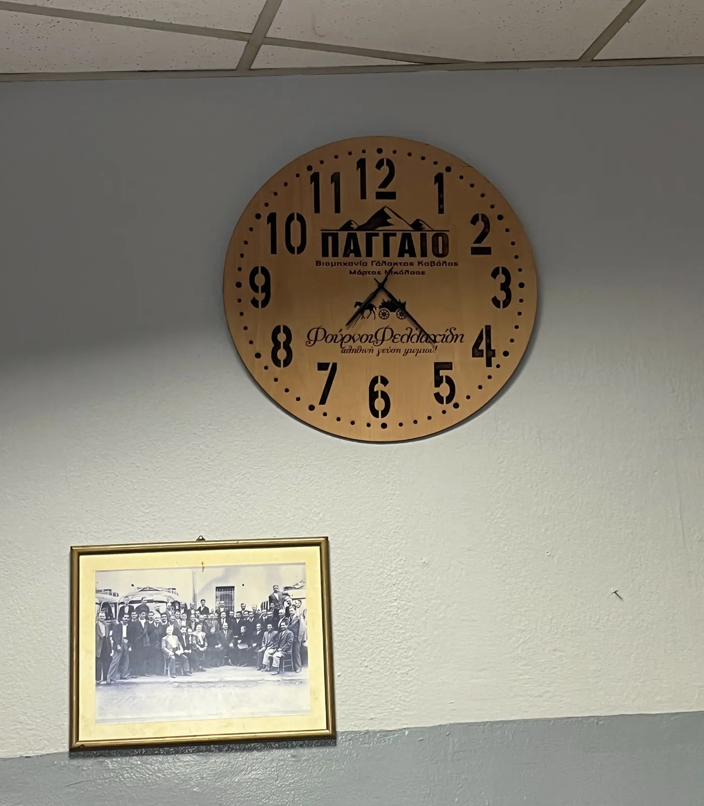 A large wooden wall clock with Greek letters on it. Next to it there’s a black and white picture of many, many people, mostly men with moustaches, standing and sitting in front of two old busses.  The clock points at 22 minutes past seven.