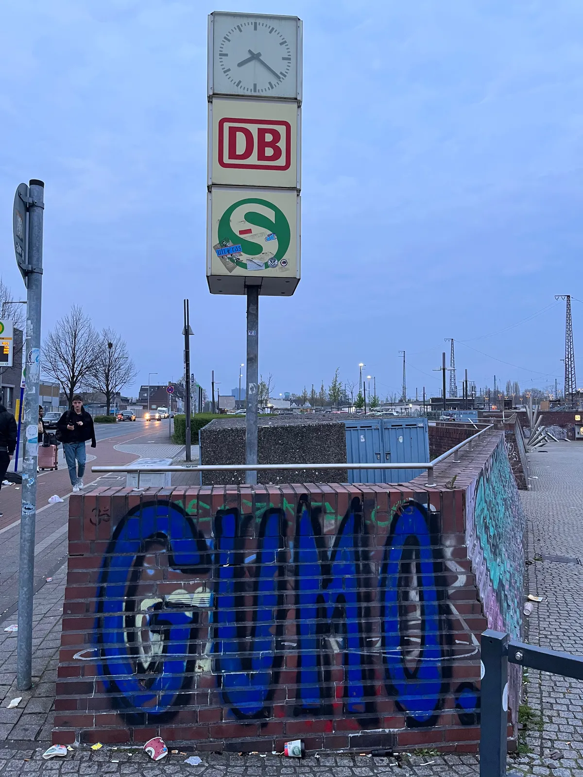 There’s a blue GUMO graffiti on a low wall. On a pole above it is a Deutsche Bahn clock. It points at 22 past 7.