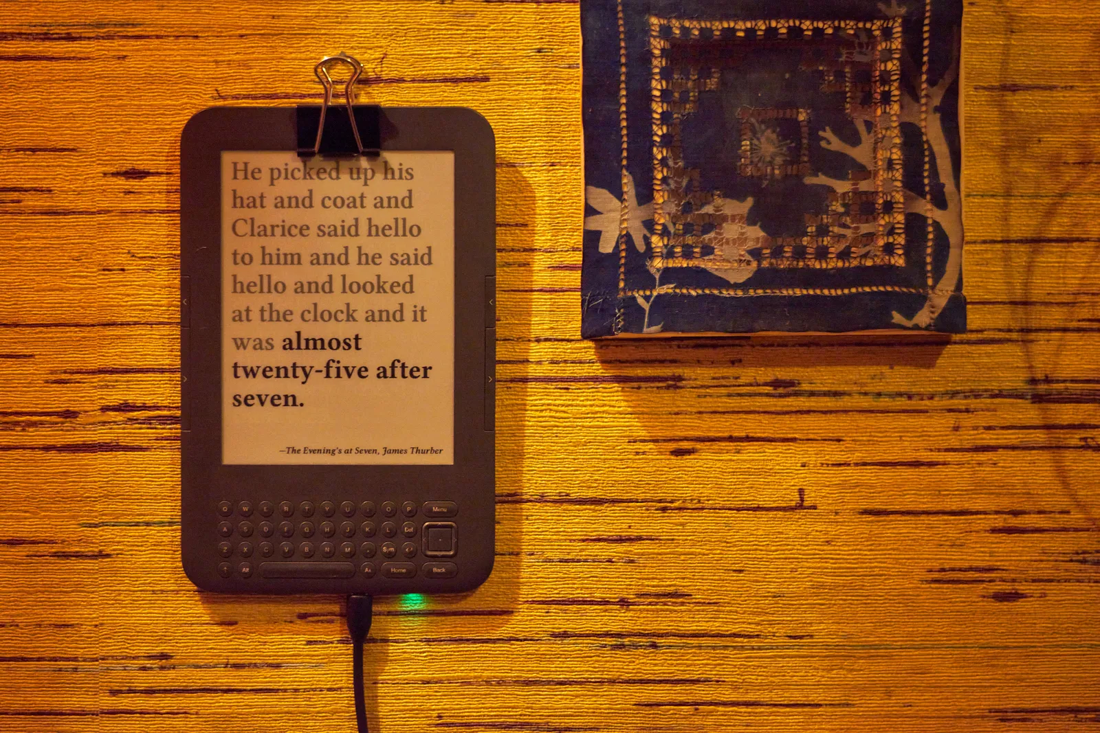 An e-reader hanging on a wall next to a piece of art. On the e-reader it says: He picked up his hat and coat and Clarice said hello to him and he said hello and looked at the clock and it was almost twenty-five after seven. -The Evening's at Seven, James Thurber