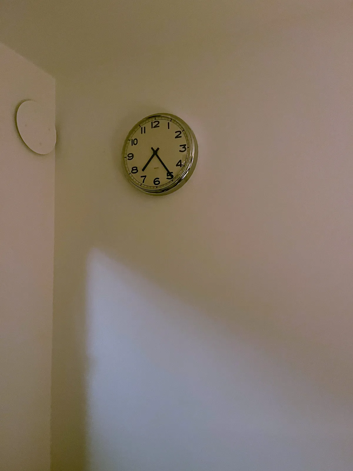 In the corner of a white room there’s on the left wall a white circle, and on the right wall there’s a wall clock that points at 24 minutes past 7.