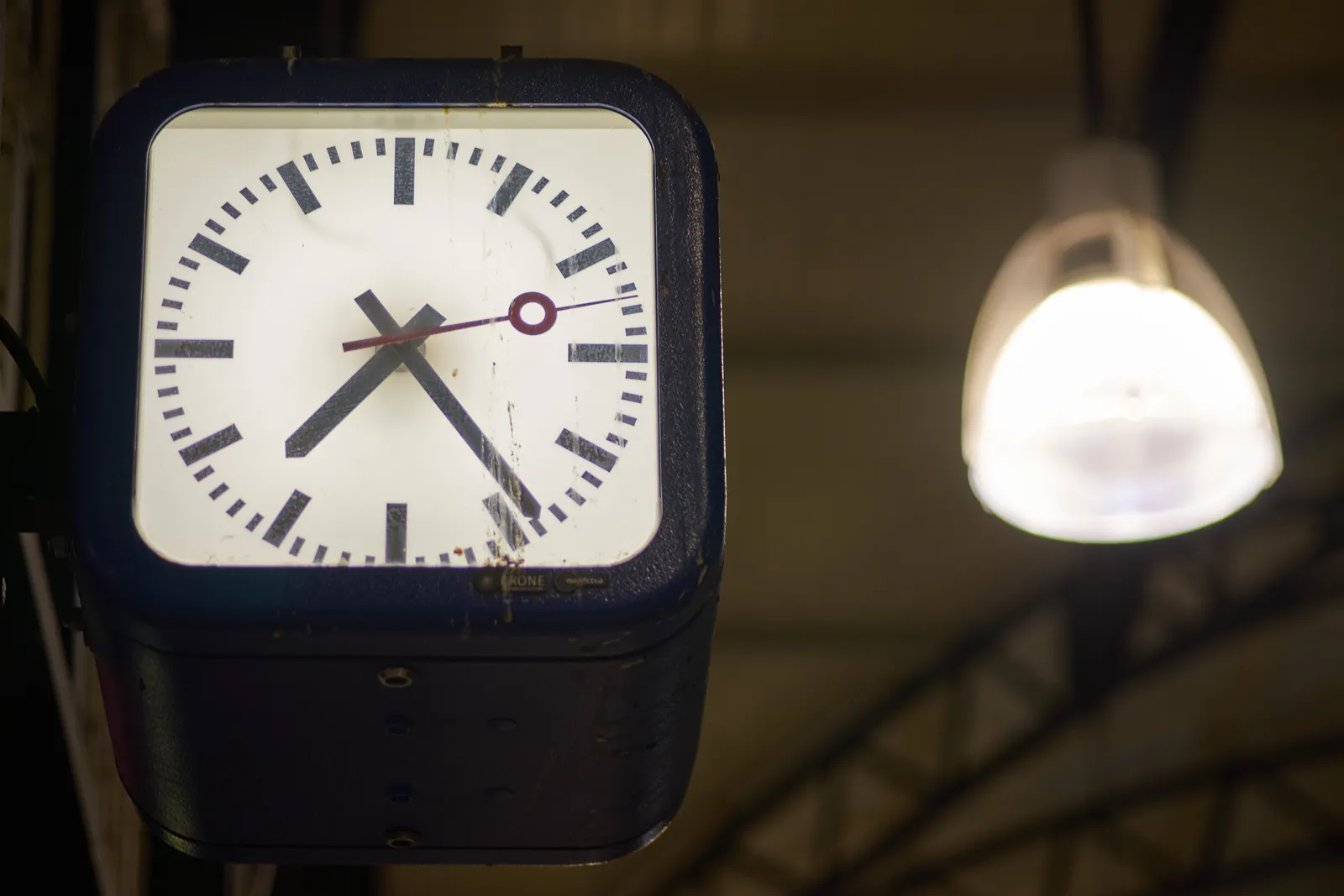 In the foreground there’s a clock that points at 24 past 7. In the background, not in focus, there’s an industrial lamp. 