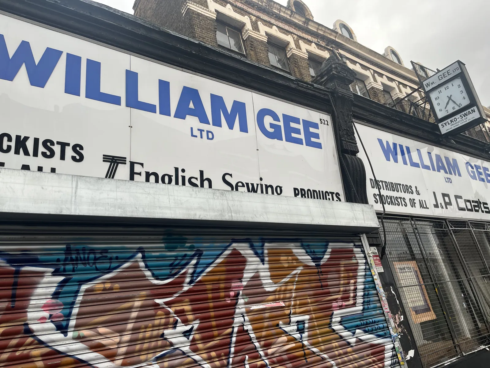 Large plastic displays on a shopping facade say that this is William Gee Limited, specialising in English Sewing products. There’s also a clock attached to the facade, which points at 26 minutes past seven. 