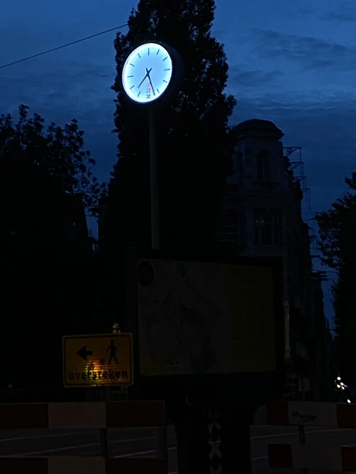 A very dark picture. Shadows of trees, shadows of buildings against a dark sky. The only thing that’s clearly lit is a clock that points at 27 past 7.