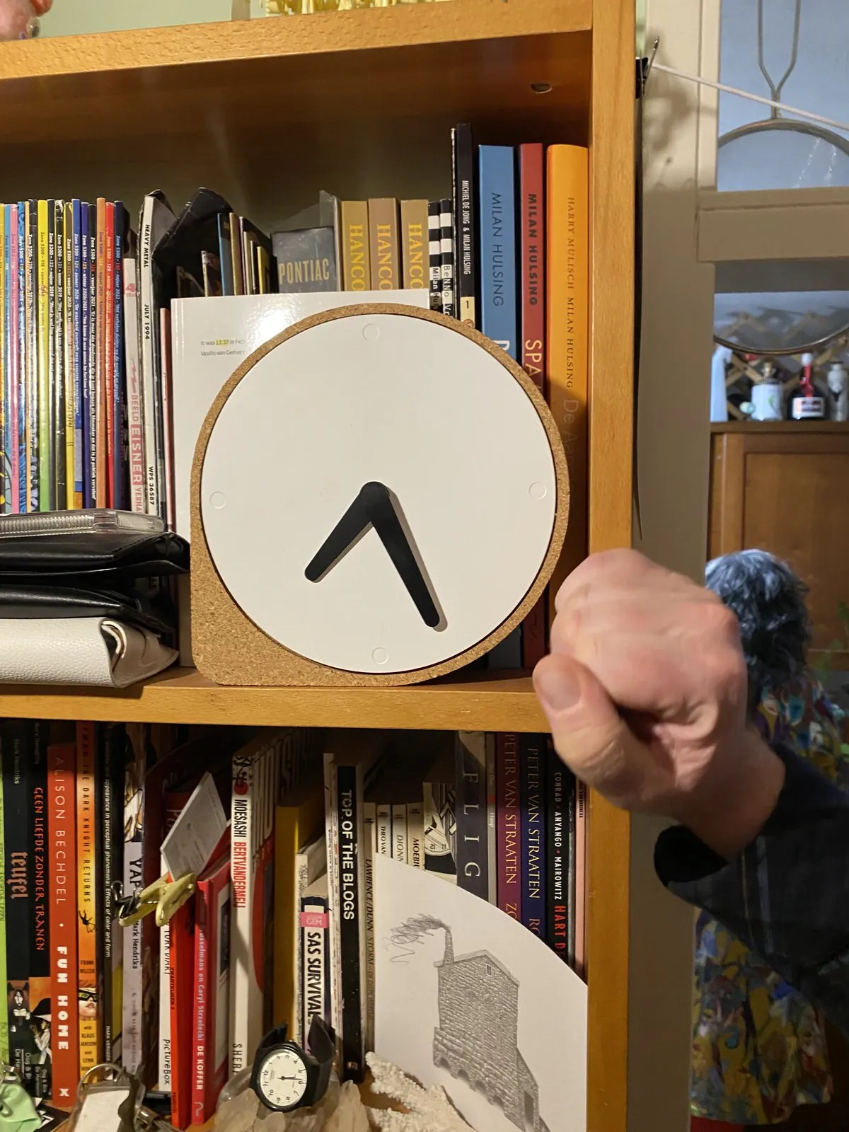 A minimalist clock in a book case points at 27 past 7. 