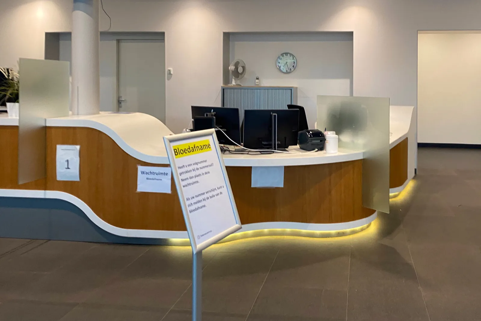 A clock behind the desk in a hospital points at 27 past 7.
