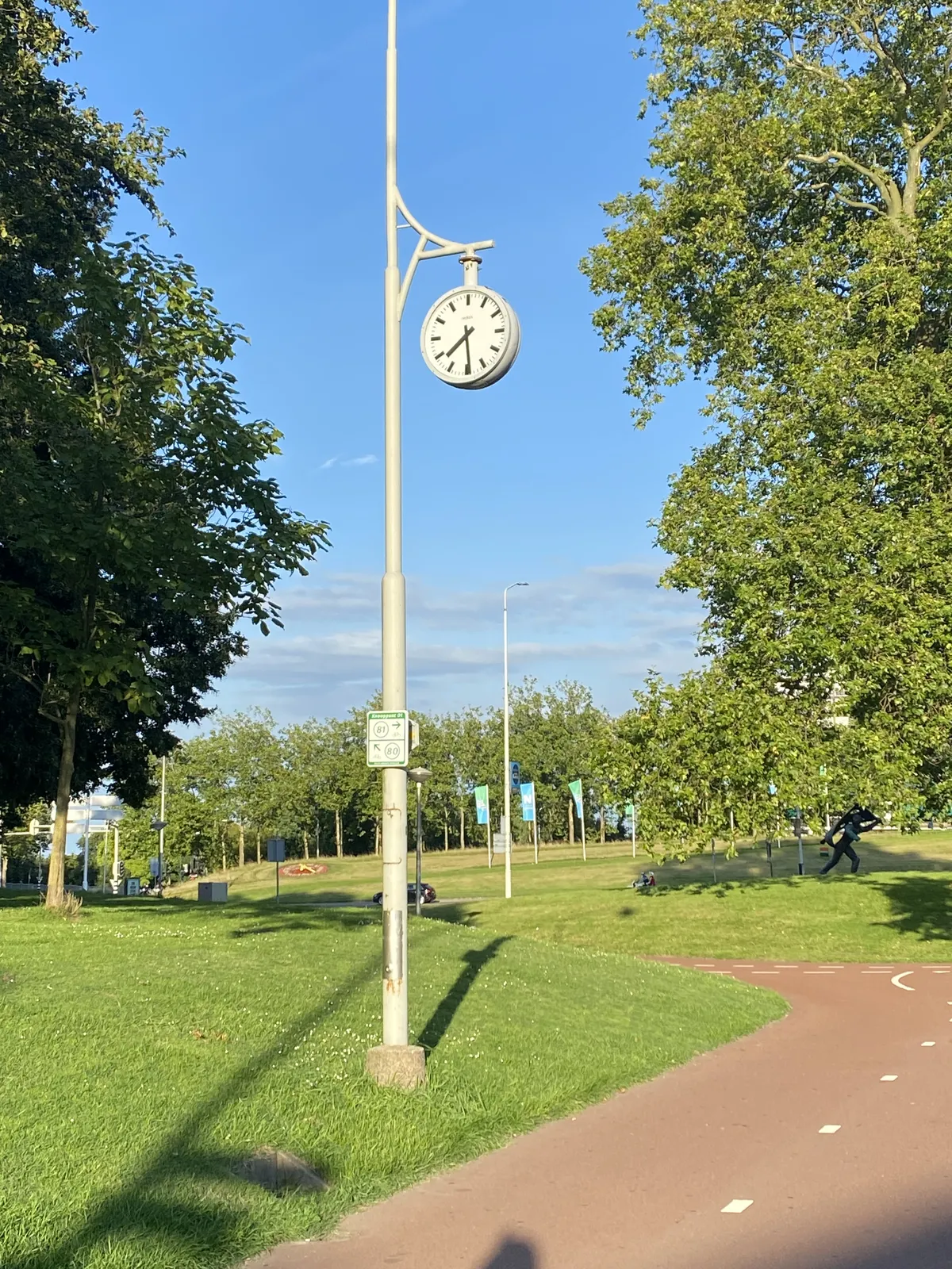 A clock hanging from a semi ornamental pole in a park. It points at 29 past 7.