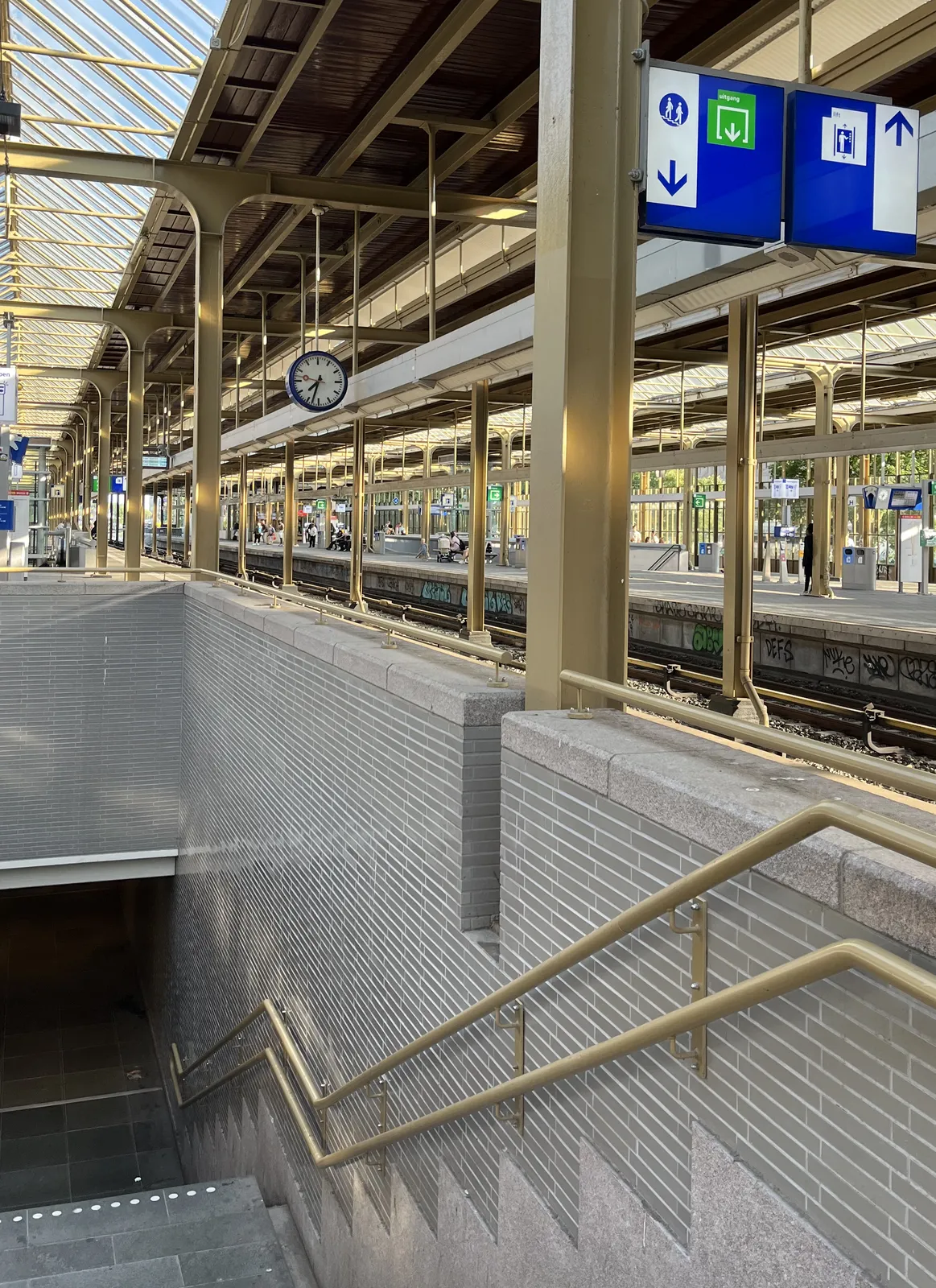 On the left of the picture there are stairs going down towards a dark tunnel. The rest of the picture is a Dutch train station. There’s a clock hanging from the ceiling which points at 33 minutes past seven.