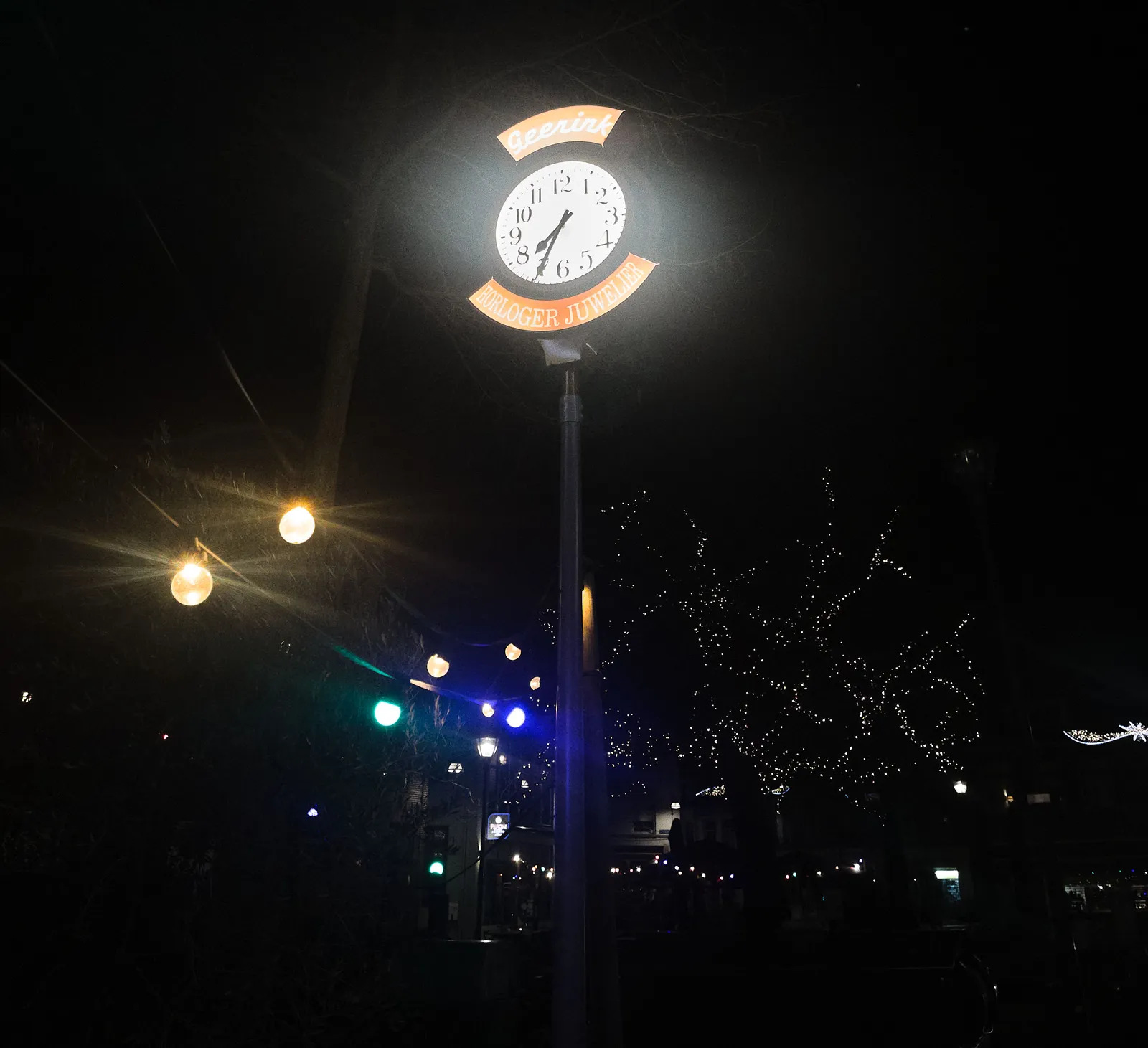 A lit clock on a pole, at night. Around the clock it says Geerink Horloger Juwelier. The clock points at 34 minutes past 7.