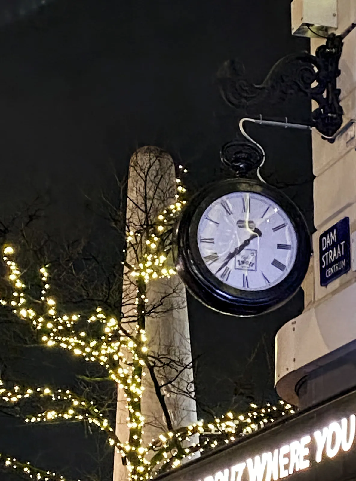 In the background the huge phallic monument of the Dam Square in Amsterdam can be seen. In the foreground, attached to a corner facade is a large clock. It points at 23 minutes to 8.
