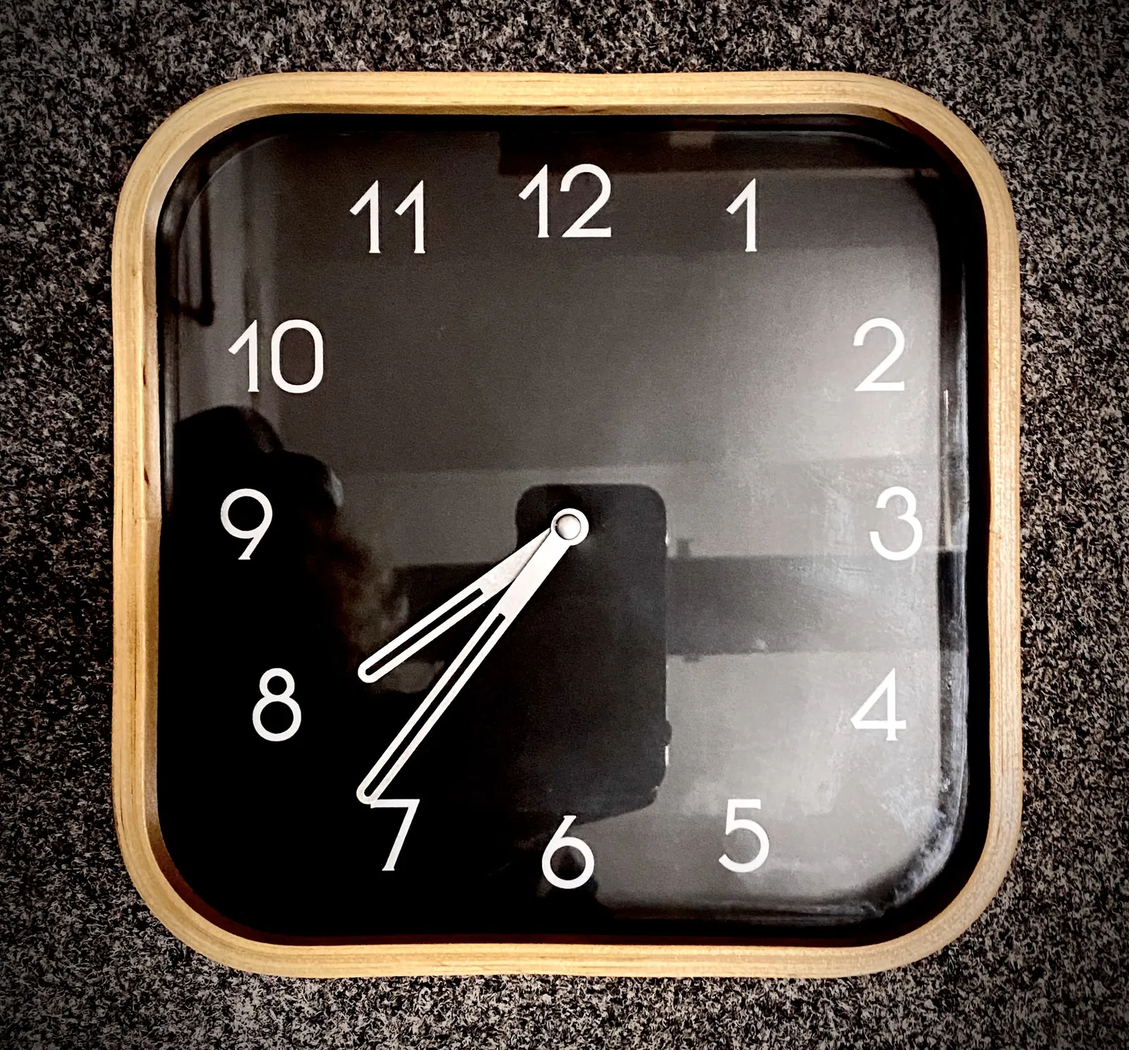 A simple, square wall clock with rounded corners. The face is black, with white numbers and hands. It points at 37 minutes past 7.