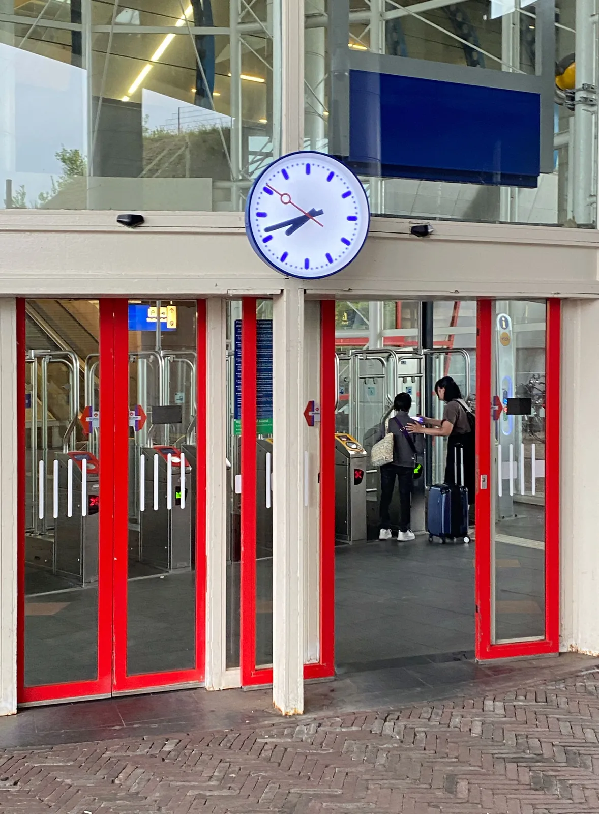 A clock on the glass facade of a train station. Through the open doors two people can be seen, the one helping the other going through the ckeck-in system. The clock points at 18 minutes to 8.
