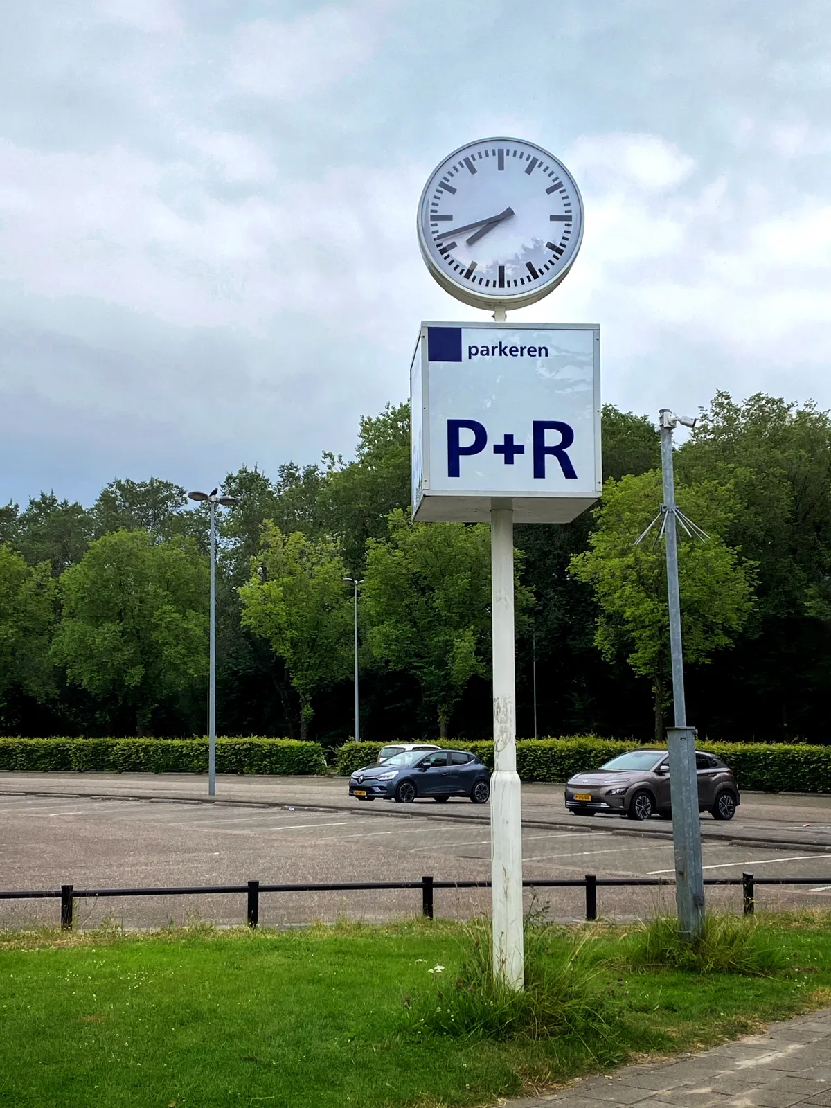 In the background there are trees, and a few parked cars. In front there’s a large sign that says that you can park here. There’s also a clock that points at 18 minutes to 8.