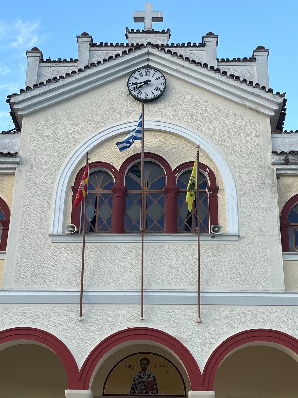 Three flags on the facade of a church. One of these flags is the Greek flag, the others are red and yellow. There’s a clock on this facade as well. It points at 17 minutes to eight.