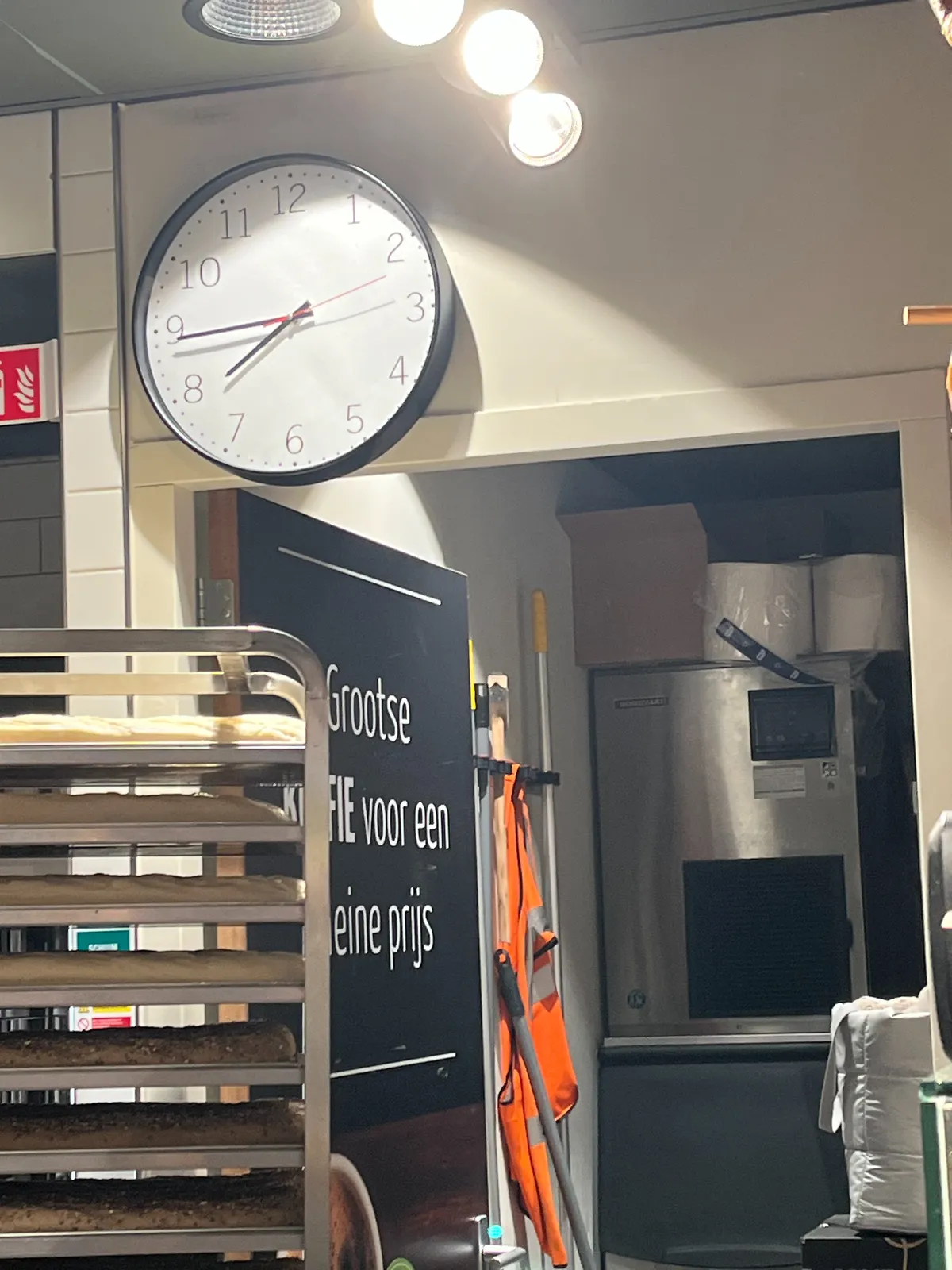 A clock hangs above a door in a bakery. There’s a trolley with trays, filled with croissants in the foreground. The clock points at 16 minutes to 8.