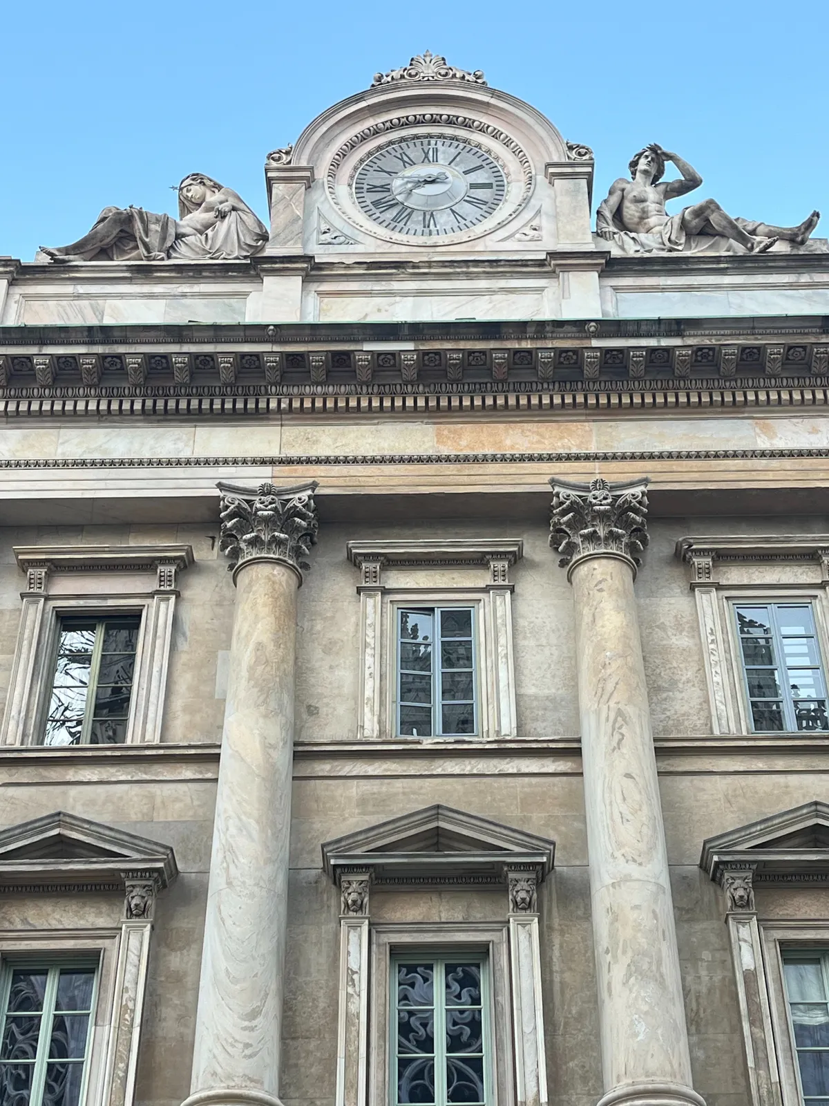 Tha facade of a large building with two Corinthian columns. On the roof there are two seated statues, a woman and a man, leaning against a clock. The clock points at a quarter to 8.