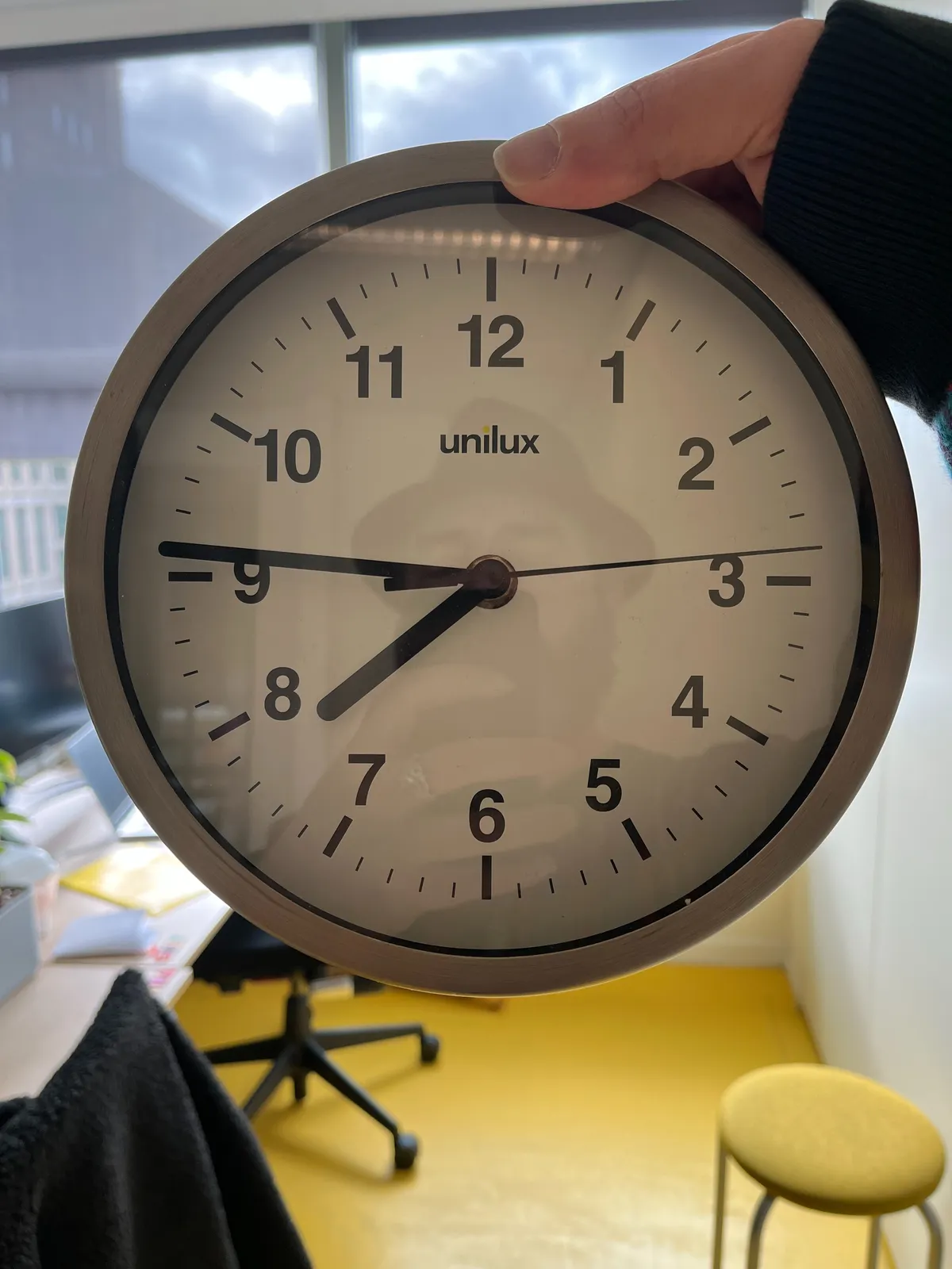 A clock is held in a hand. A man with a hat can be seen in the reflection. The time on the clock is 14 minutes to eight.