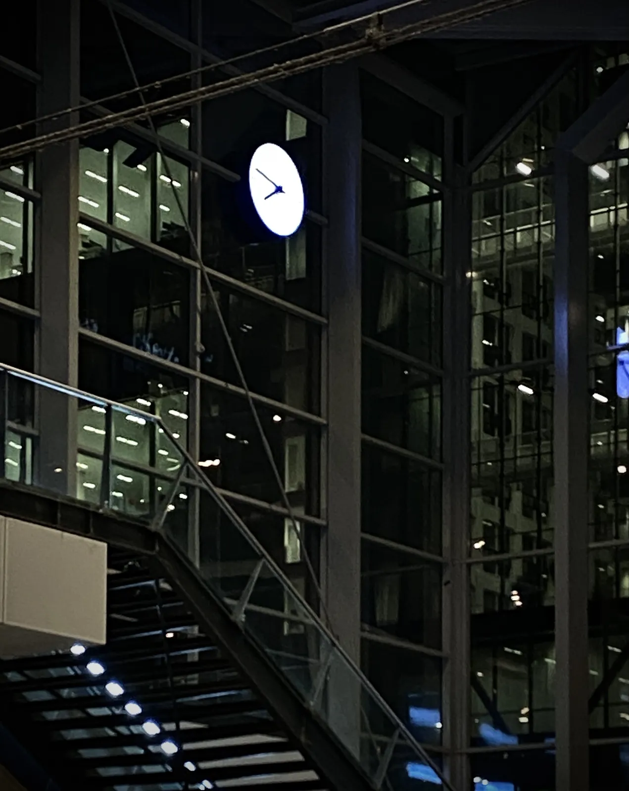 A large lit clock hanging high on a facade of a Dutch train station. It seems to point at approximately 13 minutes to 8.