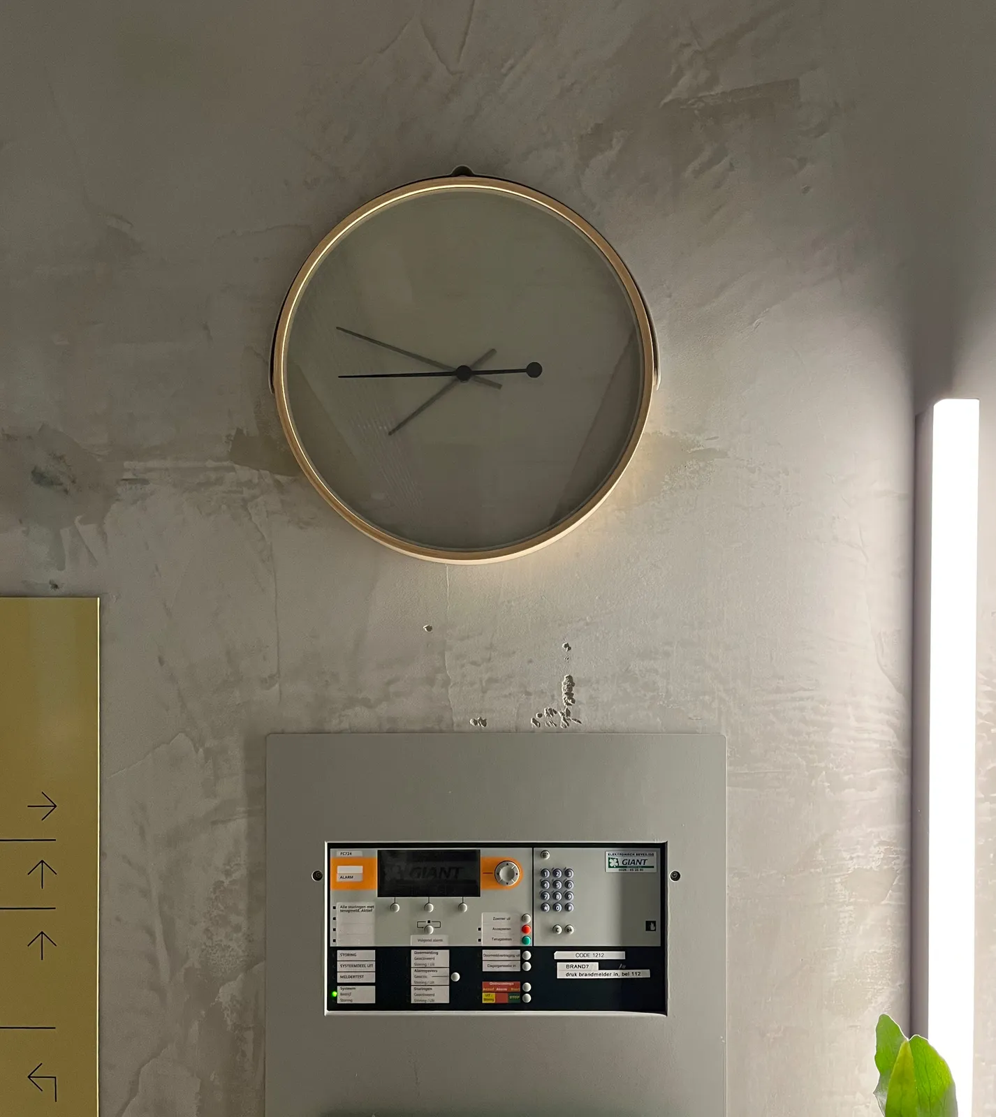 There's some sort of a control panel, a wayfinding sign, and a clock attached to a concrete wall. The clock points at 12 to 8.