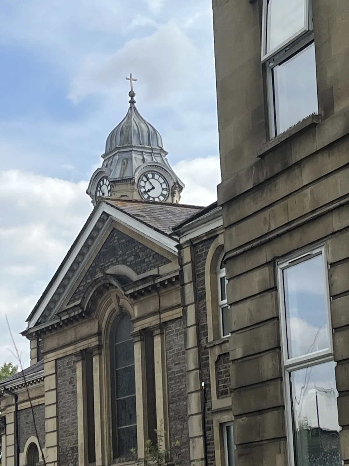A clock on a tower which peeks at us behind a brick building. The clock points at seven minutes to eight.