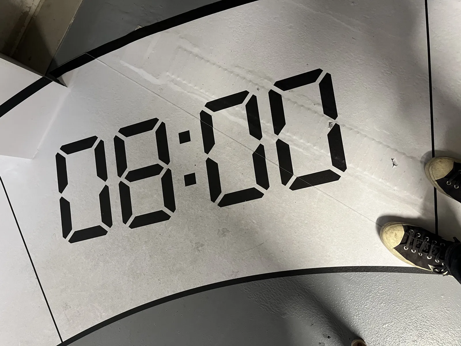 Two feet of a person standing on a floor. The timestamp 08:00 is printed in large digital digits on the floor. 