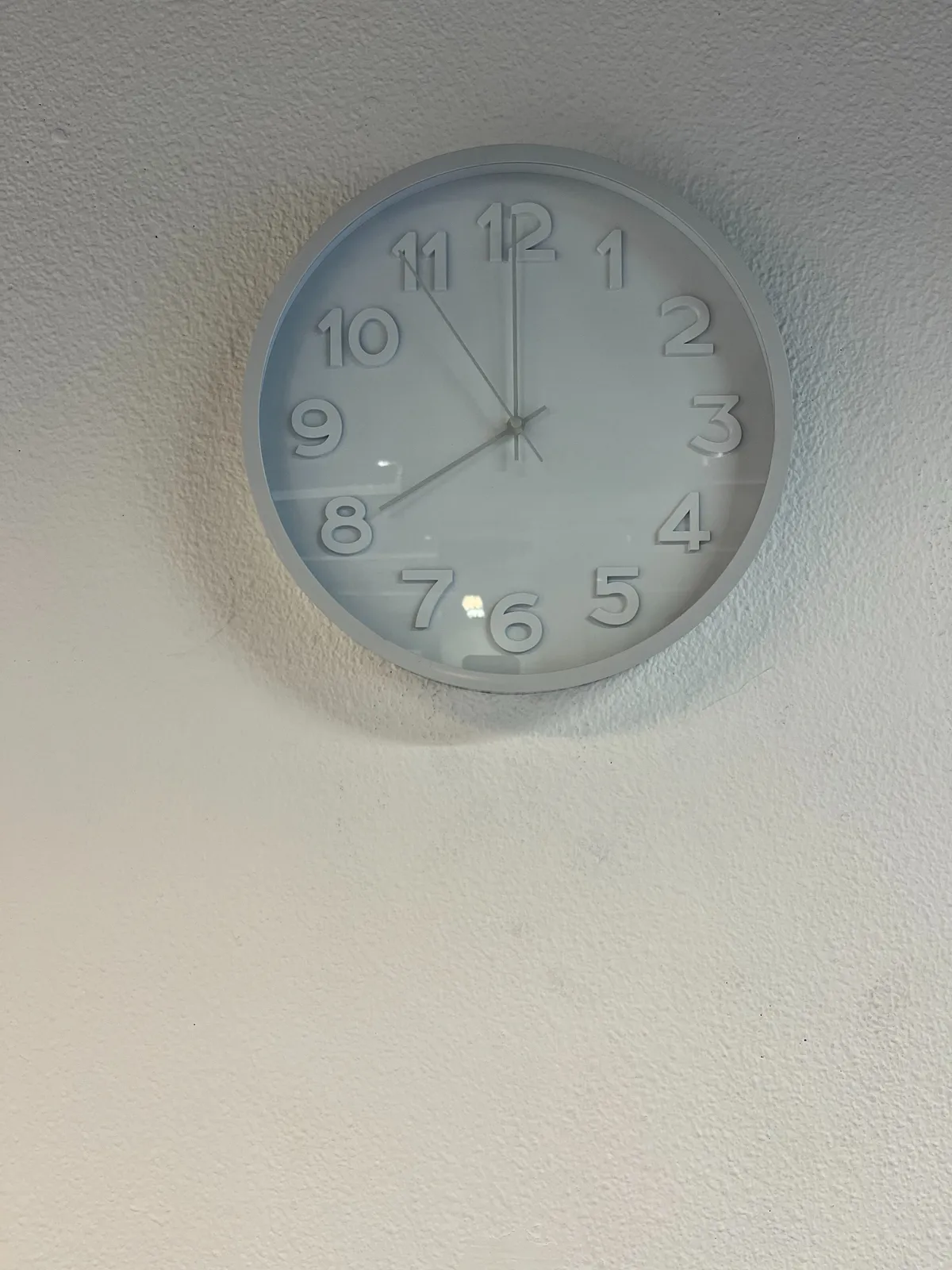 A while clock on a white wall that points at 8 o’clock.