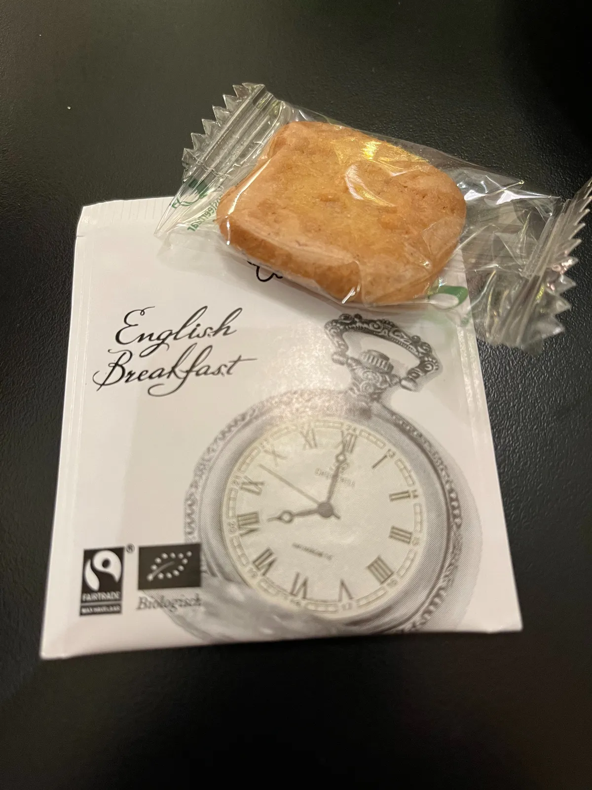 A biscuit wrapped in a piece of plastic on top of small piece of paper. Printed on this paper are the words English Breakfast and a clock. The clock points at eight o’clock.