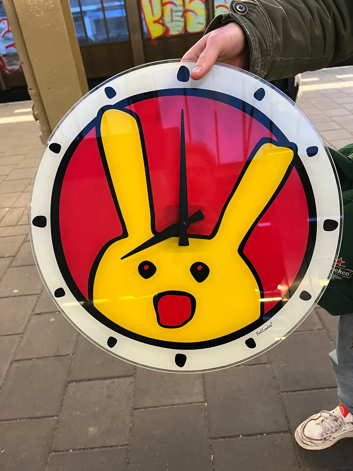 Someone is holding a brightly coloured clock with a cartoon character on it. The clock points at exactly eight o’clock. 