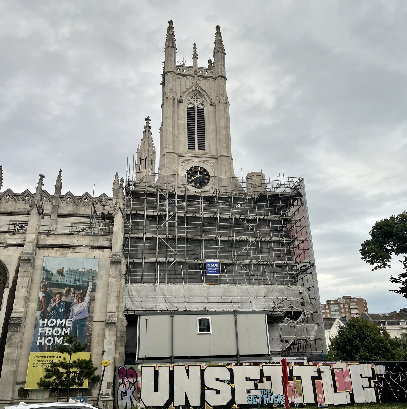 A large church, in part in scaffolding, with some large graffiti on the boards around the scaffolding. The clock on the church tower points at one minute past eight.