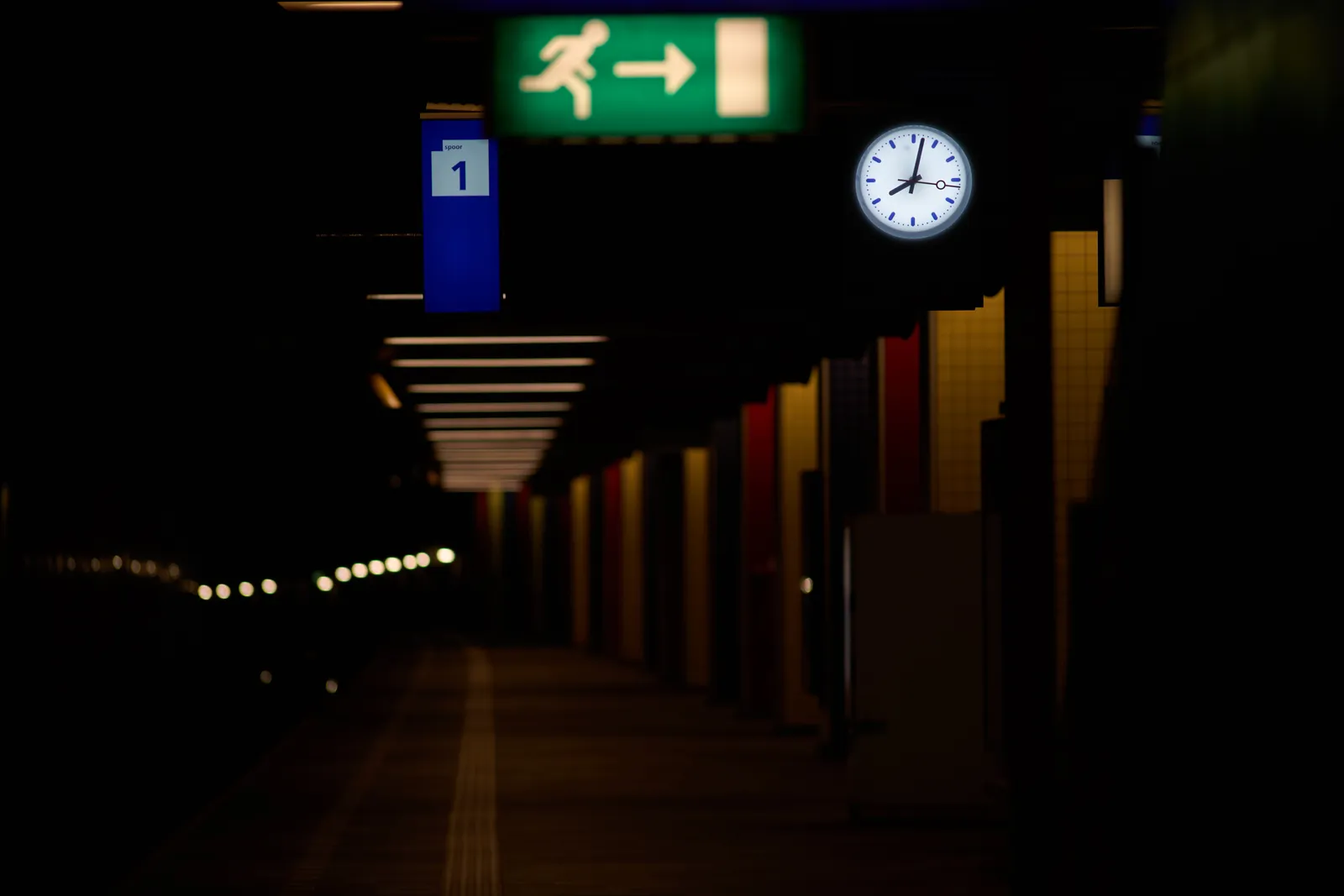 Blurry in the foreground, a green sign that points in the direction of the emerncy exit of this underground platform of a train station. There’s a clock that points at 2 minutes past 8.