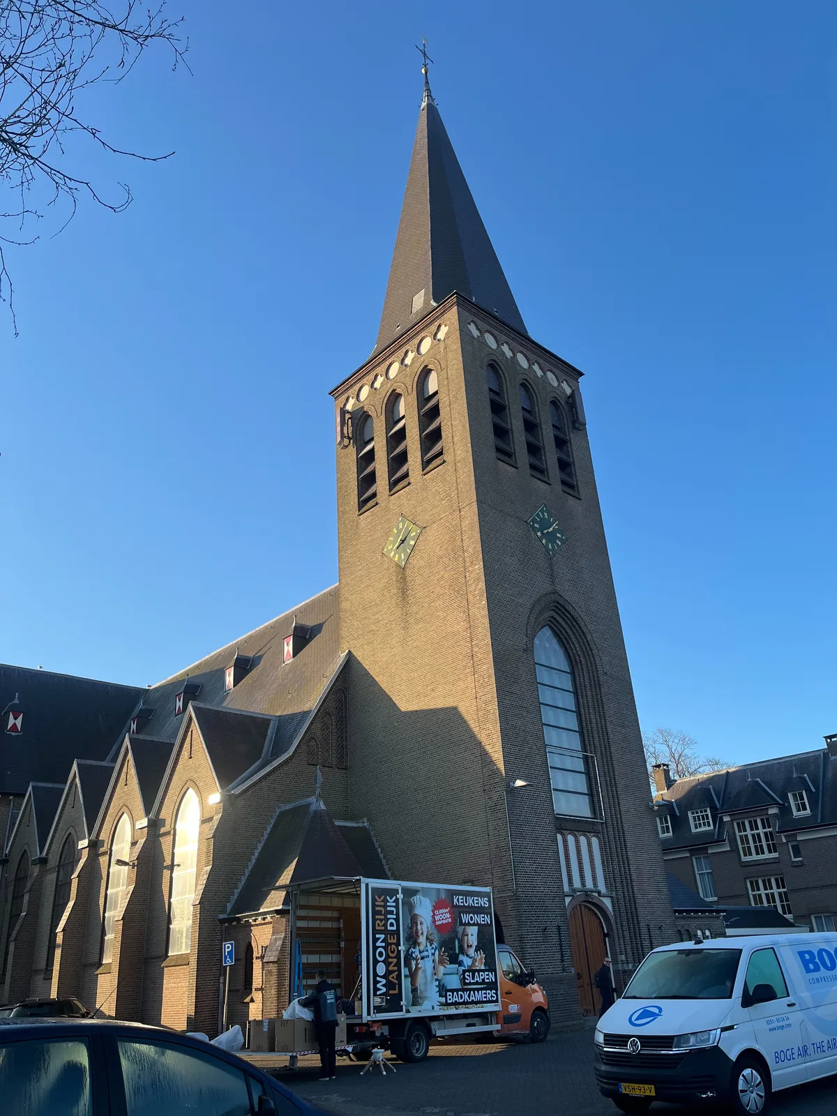 An old, Dutch, rather large, brick town church, with a few parked cars and vans parked in front of it. Two clocks can be seen on two sides of the church tower. One of them points at 5 past 8, the other at 8 minutes past 8