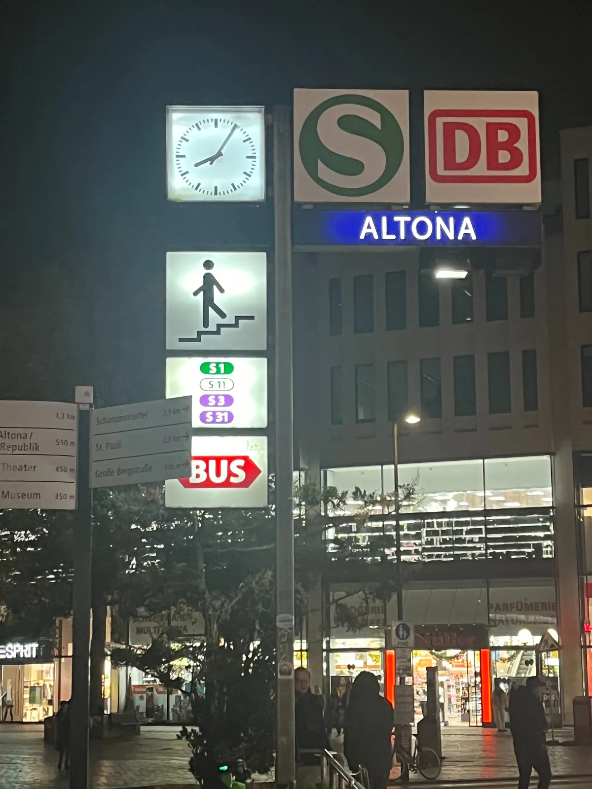 In a busy, dark cityscape there’s a sign for Altona station with a clock. The clock points at 5 minutes past 8.