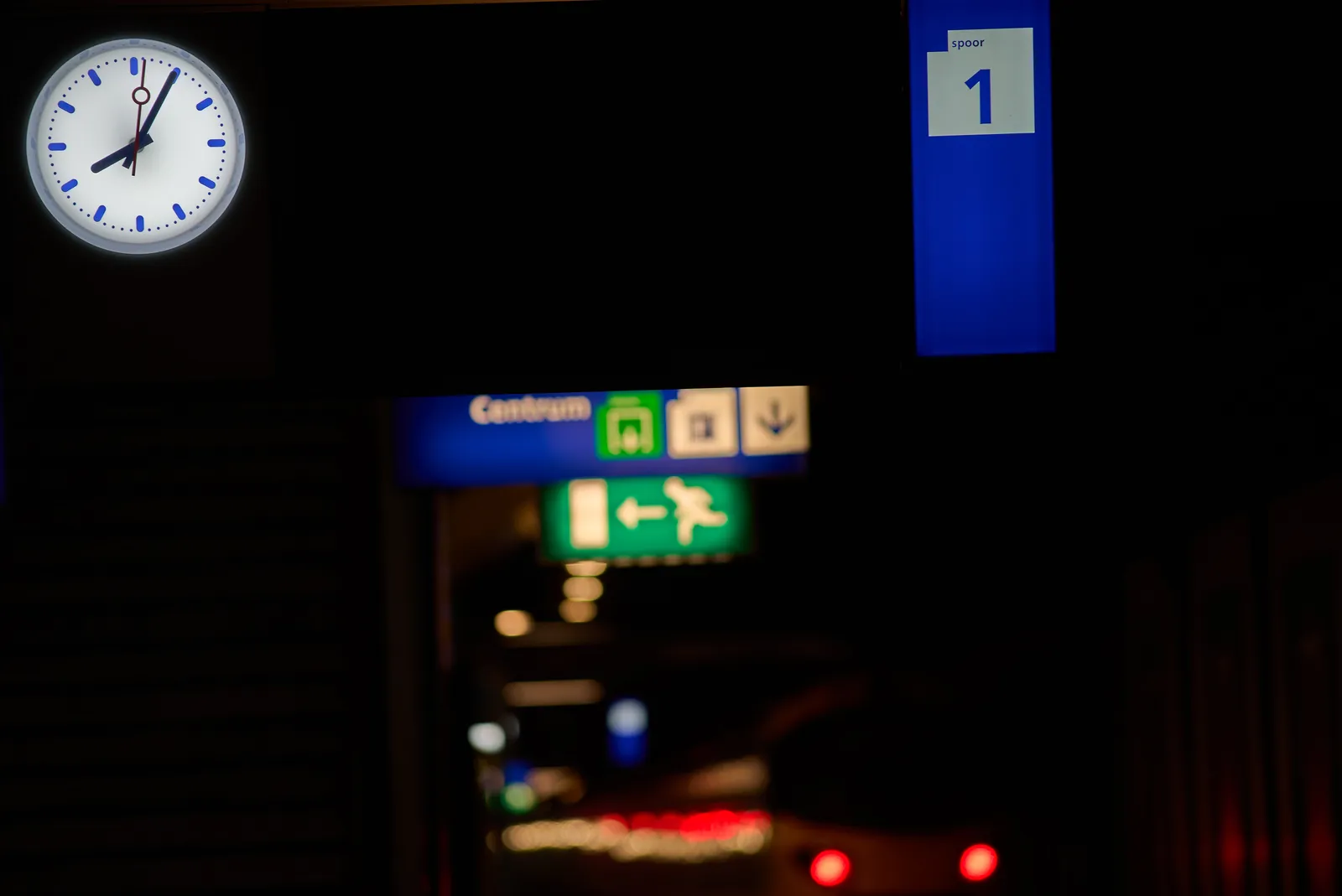 BLurry in the background a few signs and a train that’s leaving the station. In focus there’s a clock that points at 5 past 8.
