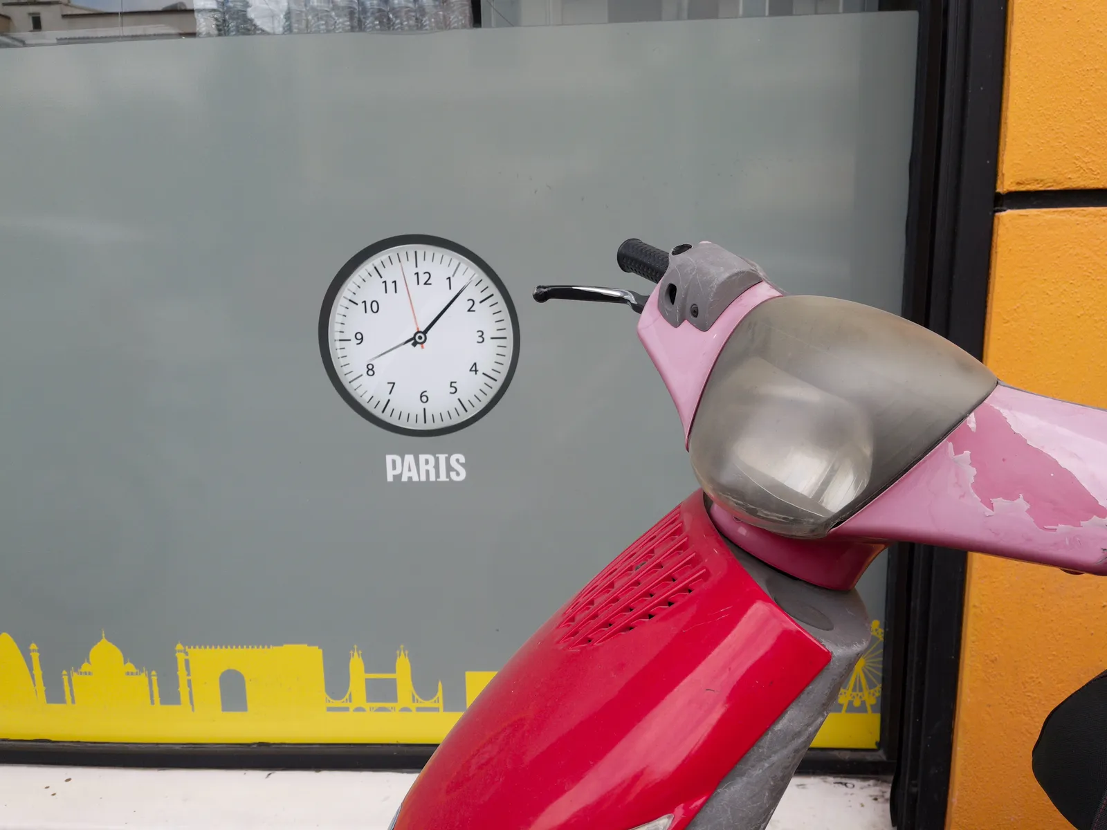 In front of a gray shopping window someone has parked their red scooter. On the window a clock is printed which pretends to show the time in Paris. Assumingly it is always 7 minutes past eight there.