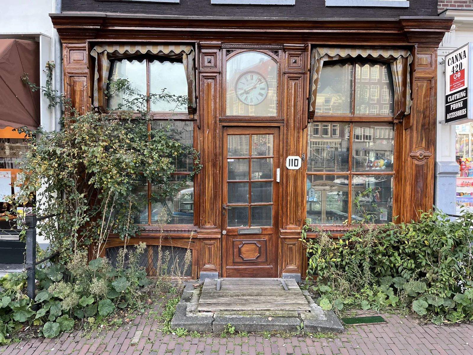 An old, wooden facade, with large green plants in front of it. The windows mirror the Amsterdam street on the opposite side of the canal. Above the door, behind glass, there’s a large clock which points at seven minutes past eight.
