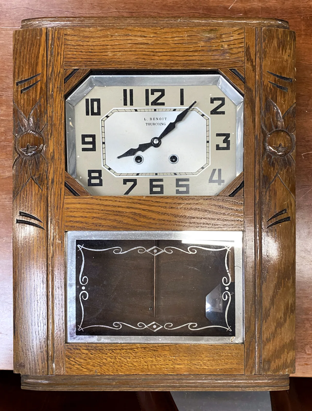 An old, wooden clock. It’s subtly ornamented, and it has a rectangular face. The numbers are rather geometrical as well. The clock points at 7 minutes past 8.