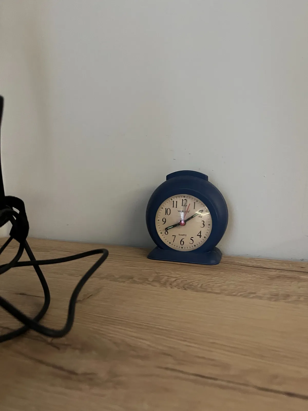 A small alarm clock on a wooden table, against a white wall. It points at 9 minutes past eight.