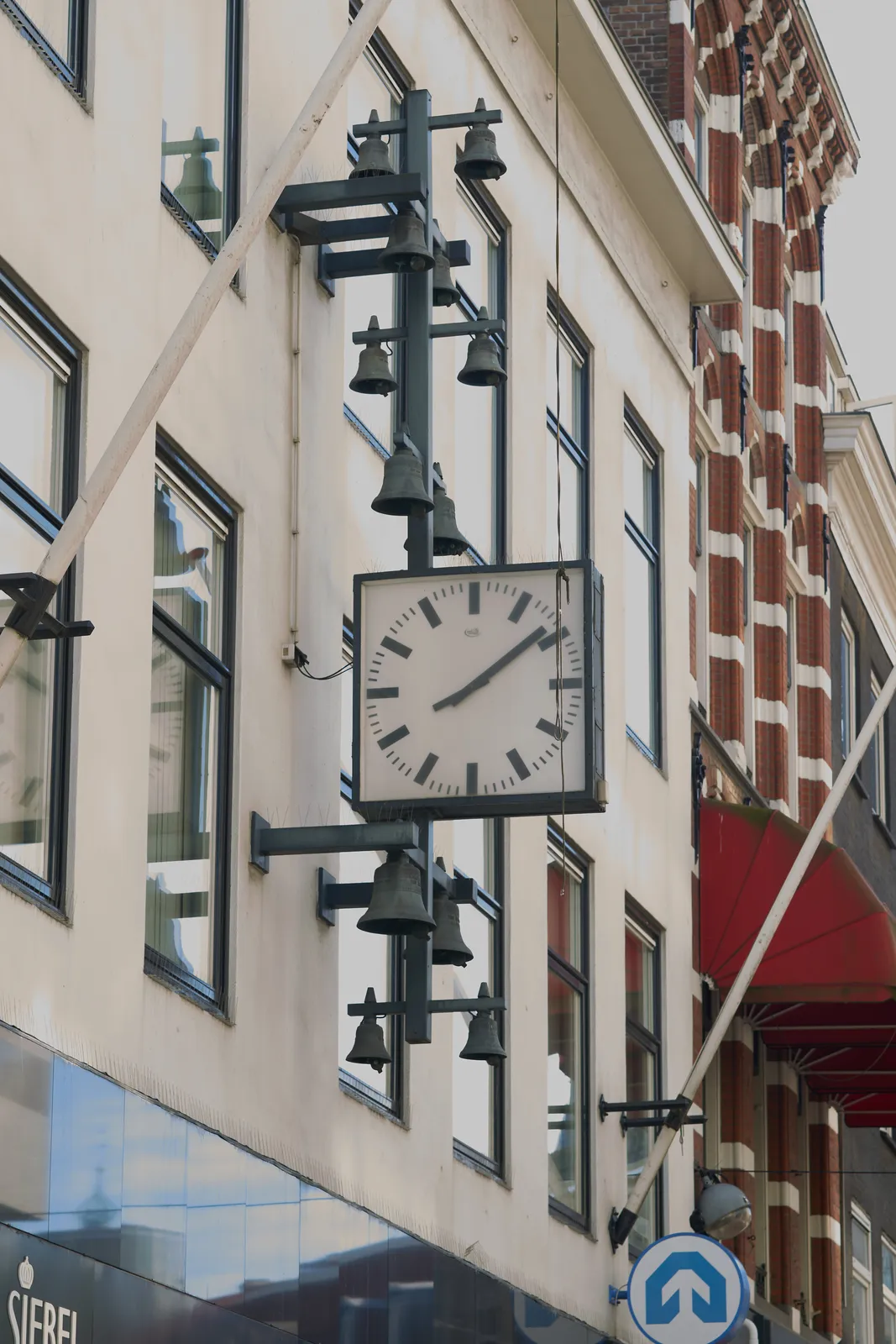 A black and white building, with attached to it a modern sculpture. It’s made from strong vertical lines, with bells, and a square clock. This clock points at 9 minutes past 8.