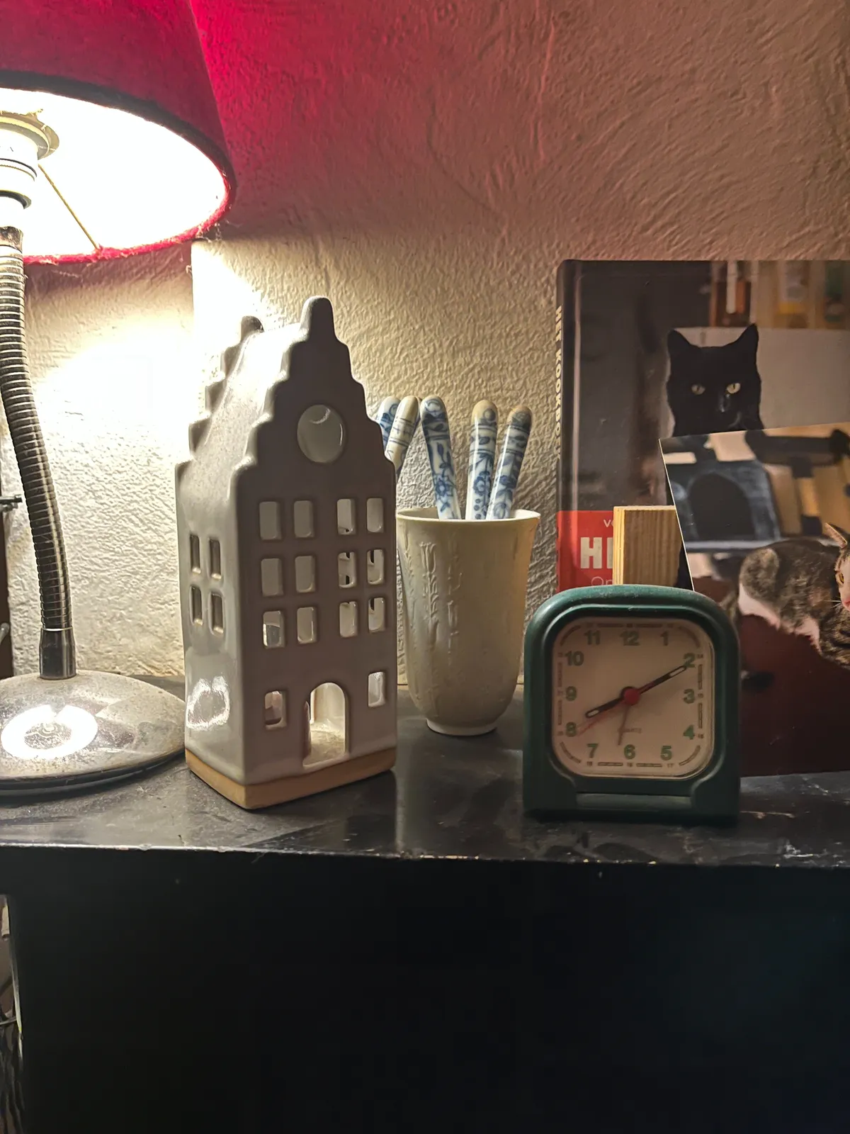A ceramic house, a table lamp, a cup with ceramic sticks in it, a few pictures of cats, and a small, analogue alarm clock. The clock points at 10 past 8.